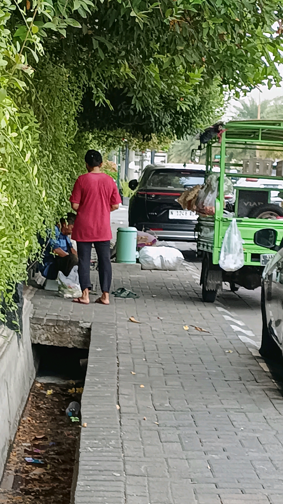 A street scene in Surabaya, Indonesia, shows a person in a red shirt and dark pants standing on a brick sidewalk. To their left, two individuals sit on the ground next to plastic bags. A green utility cart loaded with goods is parked on the right side of the sidewalk. In the background, a dark-colored car is parked. A small canal runs along the left edge of the sidewalk, containing debris. Lush green foliage overhangs the sidewalk from the left.
