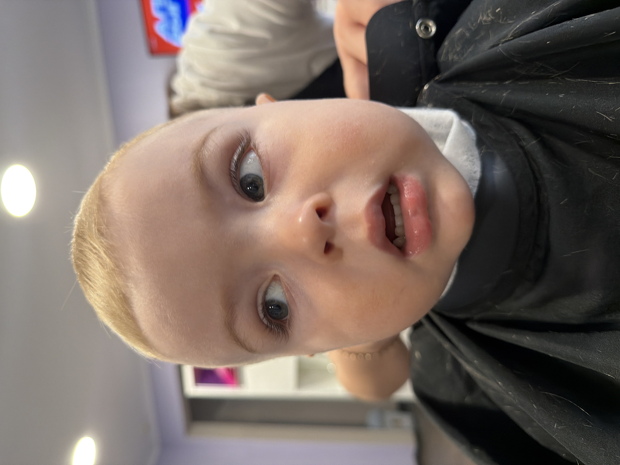 A close-up shot, oriented sideways, shows a baby wearing a black barber cape. The baby has fair skin, blonde hair, and blue eyes, and its mouth is slightly open. Scattered hair clippings are visible on the black cape. The background is out of focus, suggesting an indoor setting with purple and white walls, and a bright light source is noticeable. There is no indication that the scene is in Votkinsk, Russia.