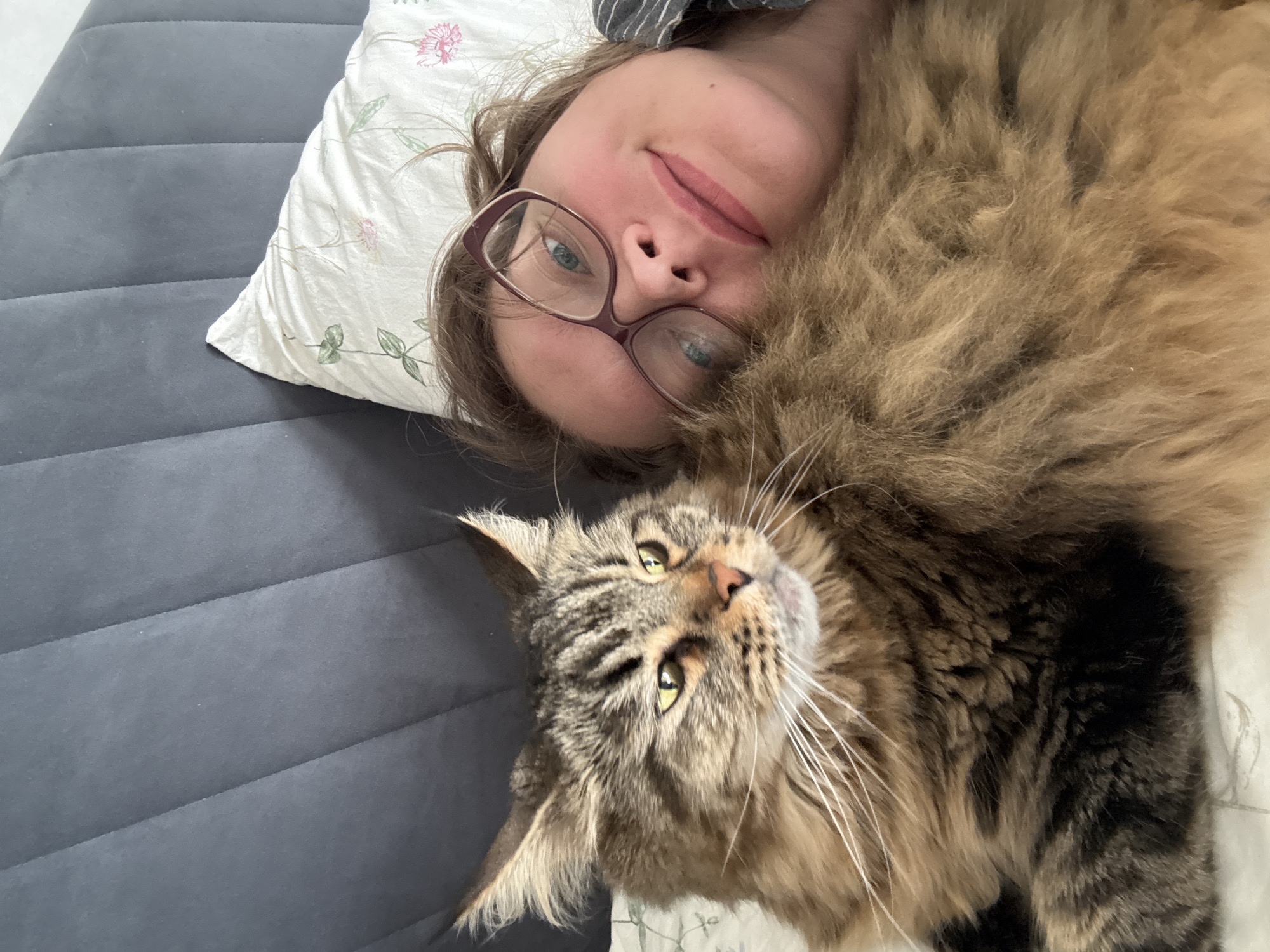 A woman with glasses is lying in bed with her cat. The cat is a large, long-haired tabby with green eyes. The woman is looking at the camera with a slight smile. The bed has a dark gray upholstered headboard and a white pillow with a floral pattern. The scene is indoors and appears to be a bedroom. There is no specific information to indicate the location is Izhevsk, Russia.