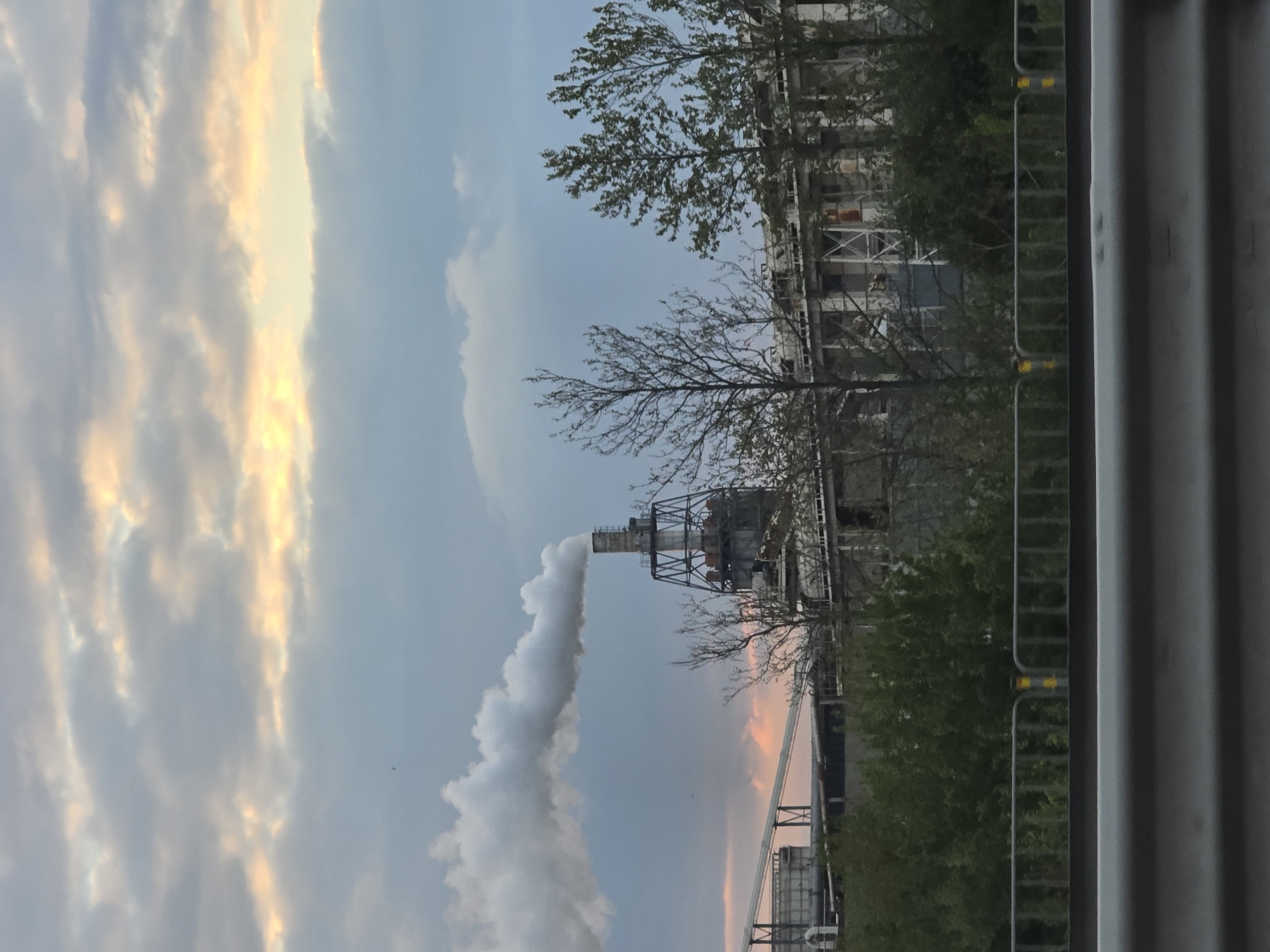 The image captures an industrial scene, likely at dawn or dusk, against a dynamic sky. In the midground, a prominent industrial facility is visible, featuring a tall chimney structure that emits a thick, white plume of smoke or steam, rising vertically and then spreading horizontally across the sky. The facility comprises various buildings and metal frameworks, suggesting a factory or power plant.
Surrounding parts of the industrial complex, and extending into the foreground, are several trees. Some trees have green foliage, while others appear more barren, suggesting a transitional season like spring or autumn. To the far right, a grey metallic barrier or fence with regular yellow markings runs vertically, partially obscuring the view and suggesting the vantage point might be from a roadside or a perimeter.
The background is dominated by the sky, which displays a mix of soft blue and dramatic clouds. Some clouds are brightly illuminated with warm golden and orange hues, particularly on the left side of the frame, indicating the presence of either a sunrise or sunset. Other clouds are darker grey, contributing to a varied atmospheric condition. The overall lighting is subdued, typical of early morning or late evening.
No people or discernible text are visible in the image. The scene is set in what is contextually known as Burgas, Bulgaria, and depicts an active industrial site amidst natural elements under a colorful sky.