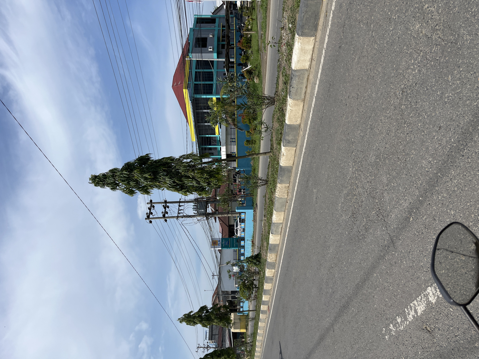 The image depicts a street scene in Tanjungkarang, Indonesia, under a partly cloudy sky. A paved road occupies the foreground, with a white and grey concrete barrier running along its edge. Beyond the barrier, a row of trees and shrubs separates the road from a blue wall.
Behind the wall, a complex of buildings is visible. One prominent building has a red, pyramid-shaped roof with yellow trim and turquoise-colored window frames and vertical accents. This building appears to be a commercial or public establishment, with air conditioning units mounted on its exterior. Another building to the left is mostly white with a blue lower section and a yellow accent. A tall, slender pine tree stands in front of this building. Further down the street, a utility pole with numerous wires is situated.
In front of the buildings, a white car is parked, and several motorcycles are visible. A sign with the acronym "PLN" in blue and white lettering is displayed on a post, suggesting a connection to the Indonesian state electricity company. The overall environment appears to be a developed urban or suburban area, with a mix of architectural styles and infrastructure. The image is taken from the perspective of someone on a vehicle, as indicated by the reflection in a rearview mirror at the bottom left of the frame.