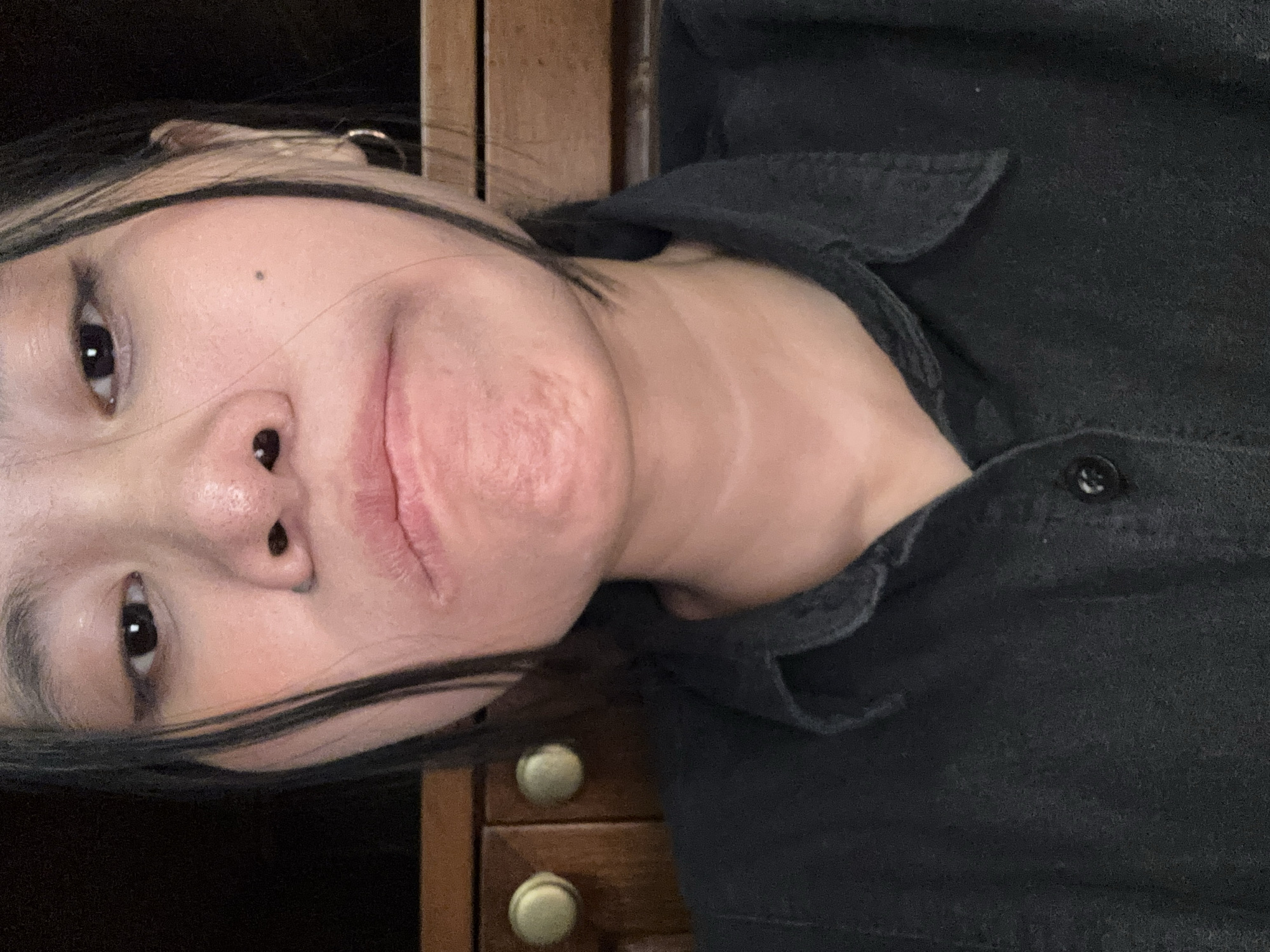 The image is a close-up, low-angle selfie of a young woman in South Jakarta, Indonesia. She has dark hair, some of which is falling around her face, and is wearing a black collared shirt. The woman's face is partially obscured by her hair, but her eyes, nose, and mouth are visible. Her expression appears to be neutral or slightly hesitant. The background consists of wooden furniture, possibly cabinets, with round knobs. The lighting is dim, creating shadows and highlighting certain features of her face. The overall tone of the image is informal and personal, suggesting it was taken in a private setting.
