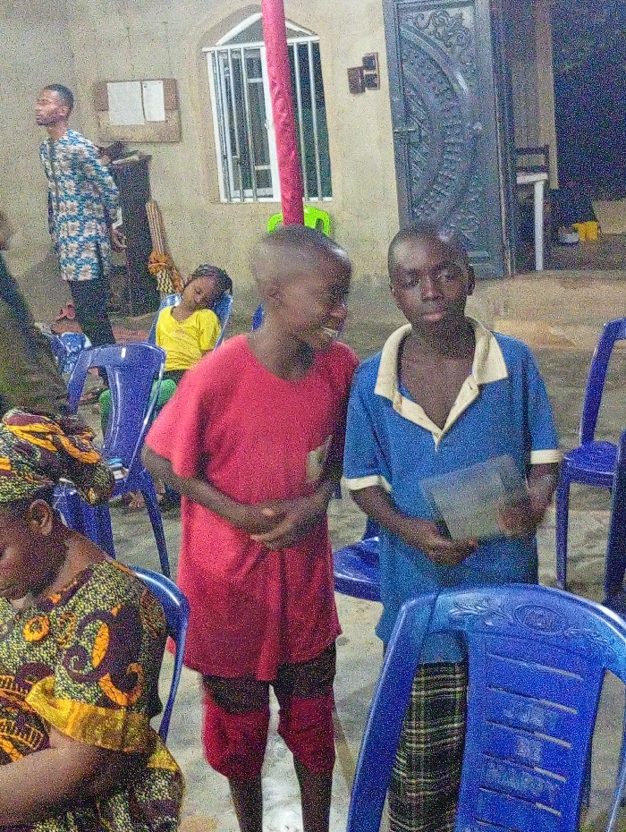 Two young boys stand close together in front of a building, surrounded by blue plastic chairs. The boy on the left wears a red shirt and matching shorts, smiling slightly. The other boy wears a blue shirt with a white collar and plaid pants, holding a dark object. In the background, a man in a patterned shirt stands to the left, and a girl sleeps in a yellow shirt. A woman in a colorful patterned head wrap sits on a chair in the foreground. A window and a door are visible behind the boys.