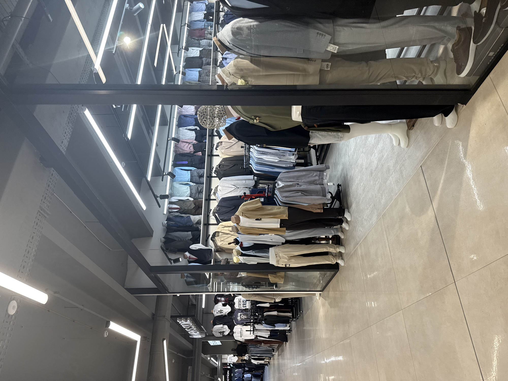 The image depicts the interior of a men's clothing store, likely in Chișinău, Moldova. The store is well-lit with fluorescent lights. In the foreground, several mannequins are displayed, dressed in casual and formal attire, including suits, shirts, and pants. The mannequins are arranged in a line, some facing forward and others sideways. Behind the mannequins, racks of clothing are visible, showcasing a variety of shirts, jackets, and pants. The store features a tiled floor and a clean, modern aesthetic. Signs for "MEN'S STORE" and other brand names are present on the walls. The overall impression is that of a typical retail environment for men's fashion.