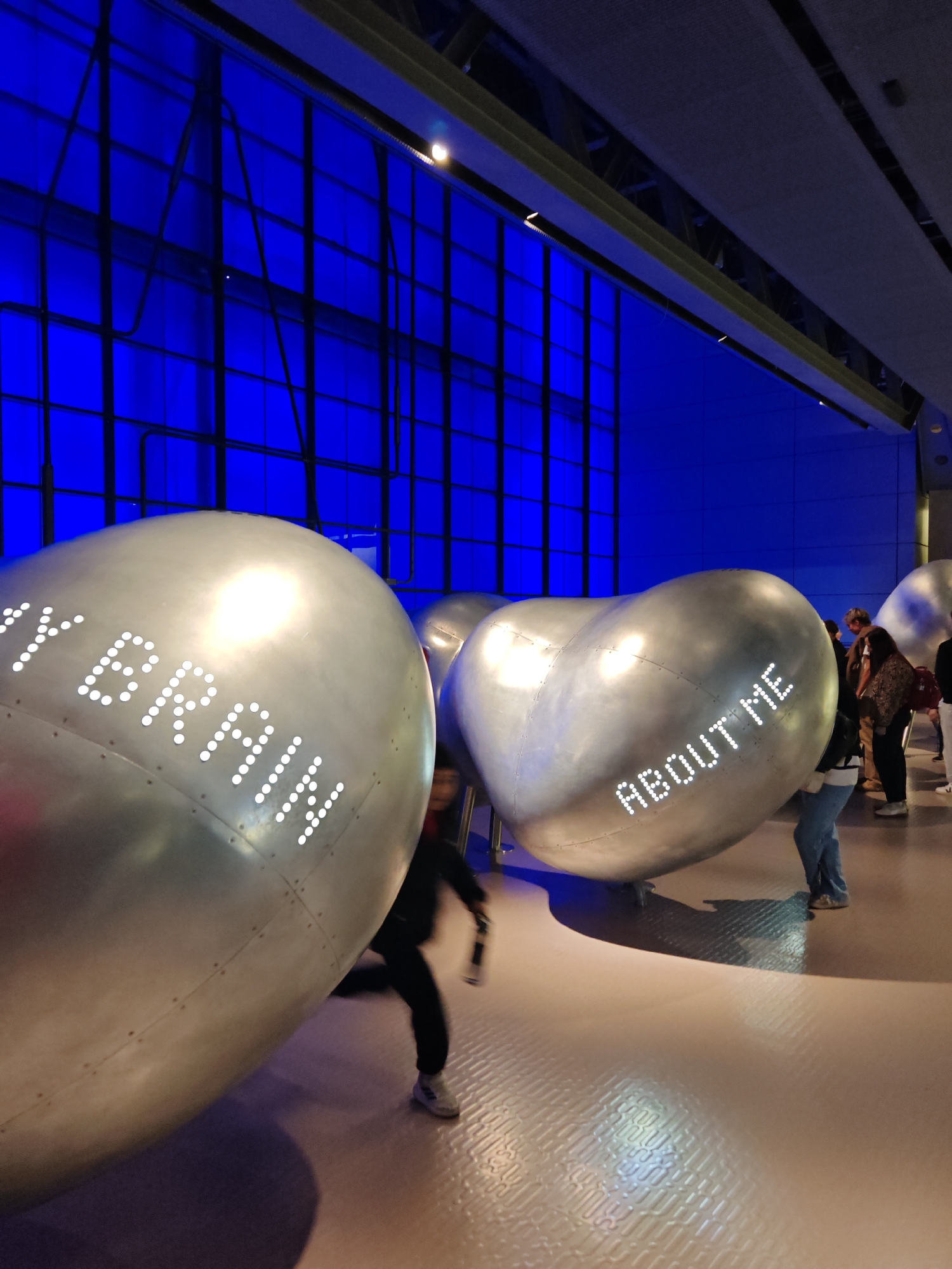 Here is a description of the image:
The image shows an interior scene at the Wellcome Collection museum in London, United Kingdom. The focus is on several large, silver, heart-shaped sculptures or art installations. The words "MY BRAIN" and "ABOUT ME" are displayed in bright, dot-matrix lettering on the surfaces of two of these sculptures. A child in dark clothing is running past the "MY BRAIN" sculpture. Several adults can be seen standing in the background, observing the display. The setting is an open area with a patterned, off-white floor. In the background, there is a wall featuring a grid of blue backlit panels. Overhead, structural beams and lighting fixtures are visible.