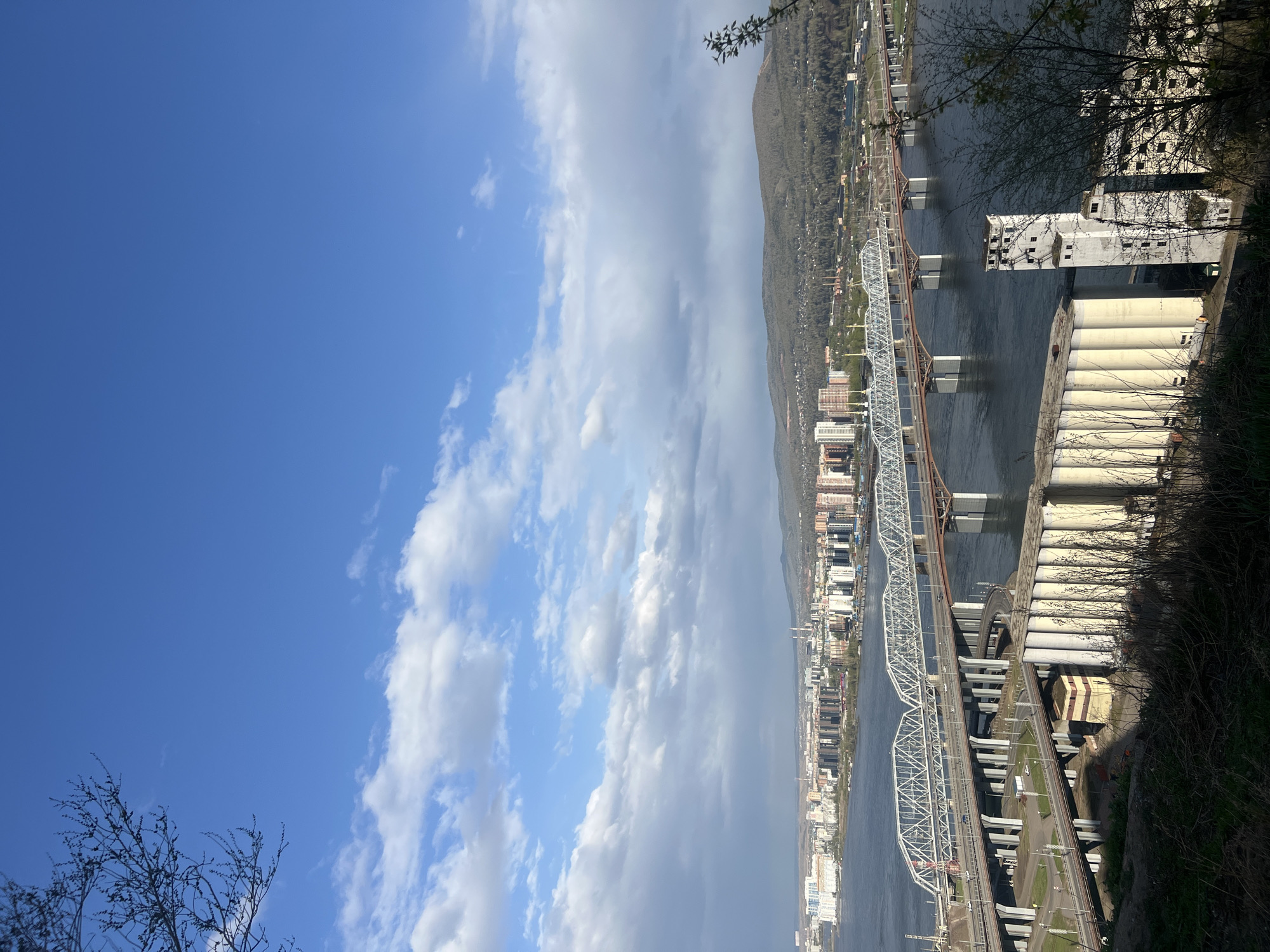 The image is an oblique, upward-facing view of a scene in Krasnoyarsk, Russia, featuring the Yenisey River, bridges, and urban structures. In the foreground is some vegetation, with bare tree branches. Adjacent to the vegetation is a cluster of silos and a building with a square, block-like structure. Moving across the river are two bridges, one painted white and the other, a rust color. On the far side of the Yenisey is the city of Krasnoyarsk, with apartment buildings and other structures in the urban setting. Behind the buildings are the mountains that surround the city of Krasnoyarsk. Above, the sky is divided between patches of clear blue and areas filled with fluffy white clouds.