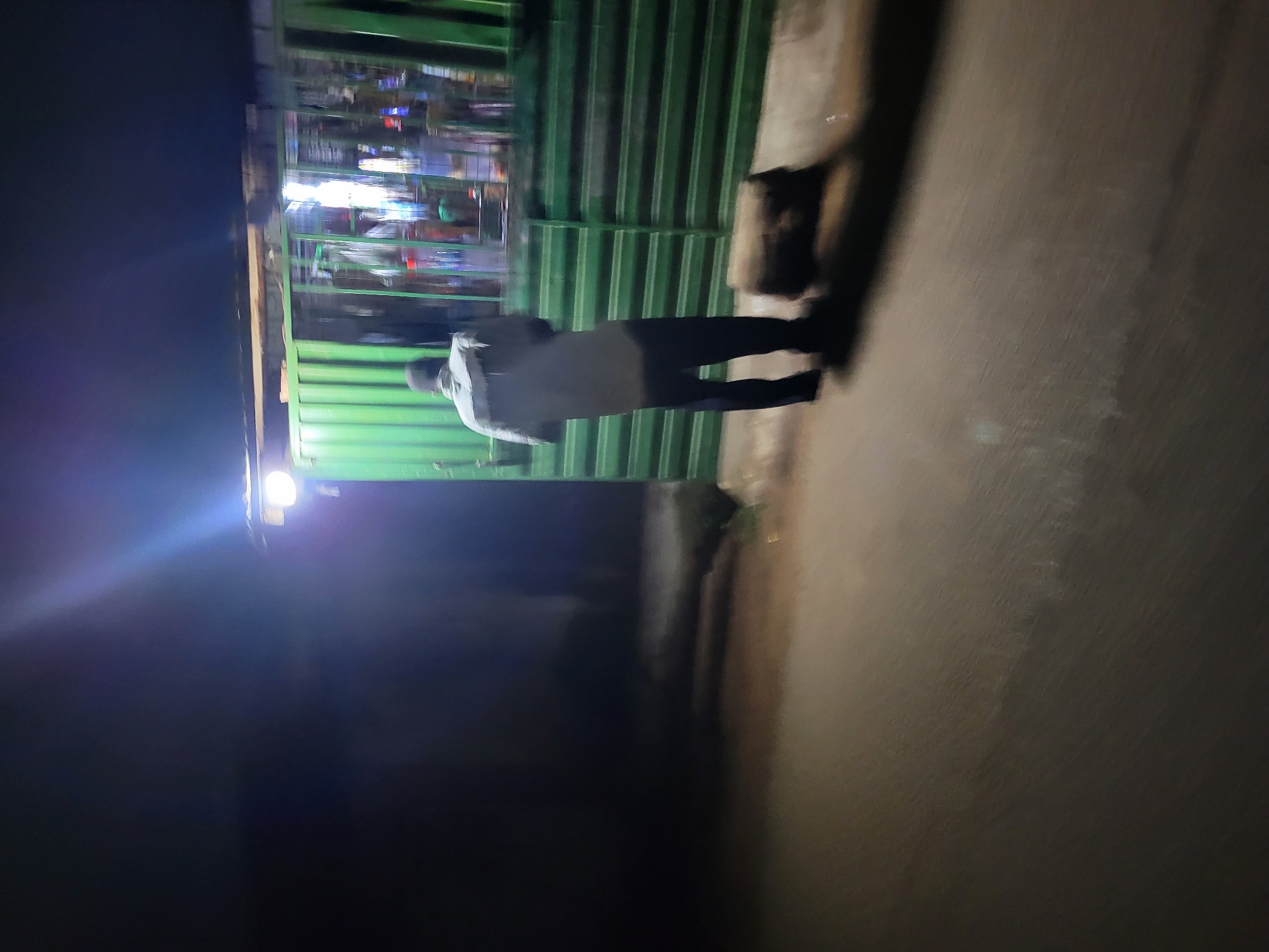 A dimly lit outdoor scene shows a man standing in front of a green, corrugated metal structure. The structure appears to be a small shop or kiosk, with visible lights and merchandise inside. The man is wearing a light-colored shirt and dark pants. He is facing the shop. The ground appears to be paved. The scene is dark, suggesting it is nighttime. The location is the city of Jos, Nigeria.