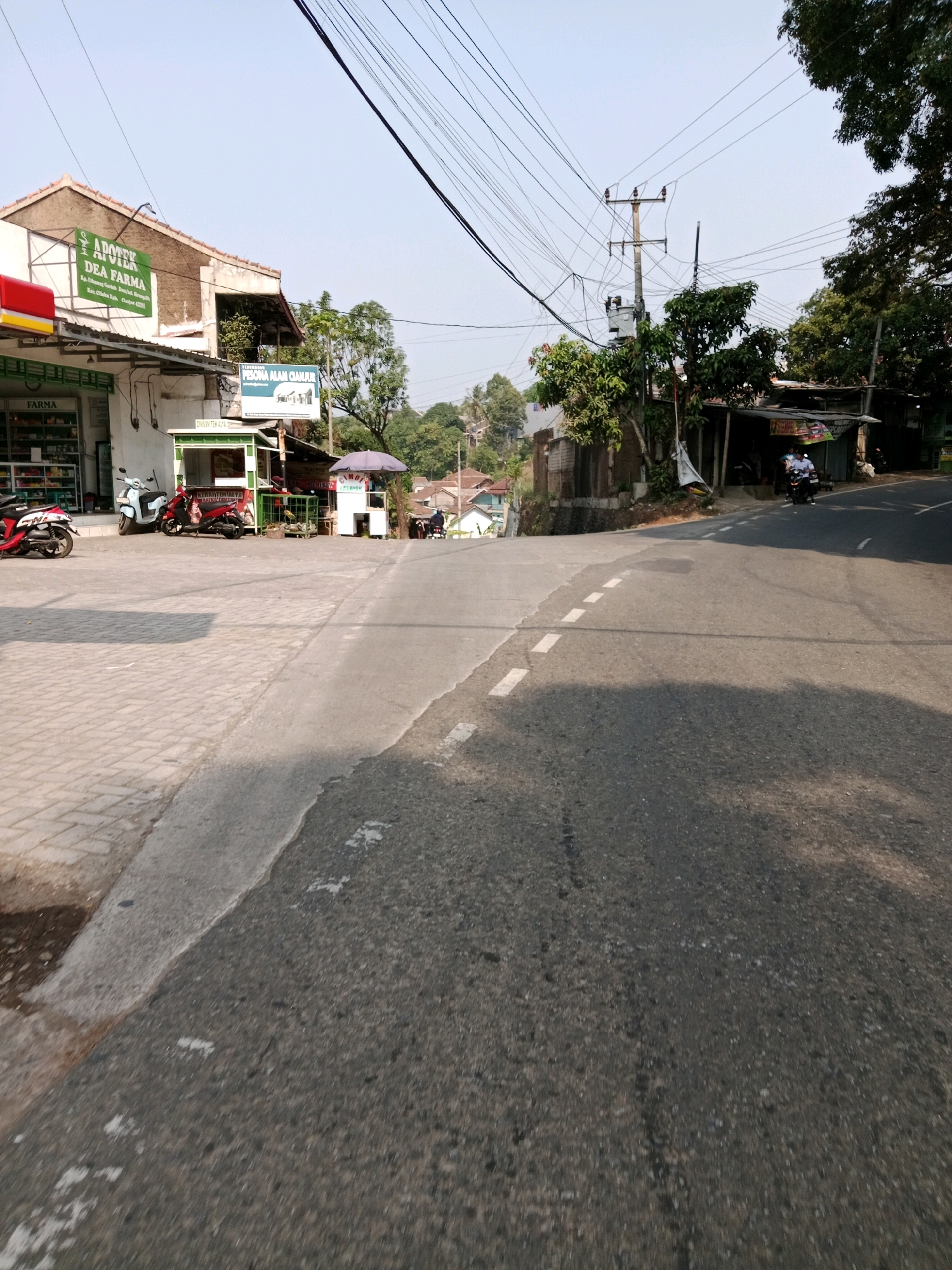 The image depicts a street scene in Cilaku, Indonesia, on a clear day. A paved road with a broken white line marking runs diagonally from the bottom right corner upwards towards the left. The road is bordered on the left by a sidewalk made of interlocking concrete pavers.
Along the sidewalk, a row of commercial establishments is visible. The most prominent is a building with a sign that reads "APOTEK DEA FARMA" in green lettering on a white background. Below this sign, another sign indicates "FARMA" in red lettering. To the right of the pharmacy, there's a small green kiosk with the words "DIMSUM TEH ALAMI" visible. Further to the right, a small white stall with a purple umbrella over it displays a sign that appears to read "GAMOI."
Several motorcycles are parked along the sidewalk. A red and white motorcycle is parked on the left, a light blue scooter is next to it, and a red motorcycle is parked in front of the green kiosk. In the distance, on the right side of the road, a person on a motorcycle is riding away from the camera.
The environment includes a power pole with numerous electrical wires crisscrossing overhead, and lush green trees are present throughout the scene, some obscuring parts of the buildings and the sky. In the background, residential buildings can be seen on a gentle incline. The overall impression is that of a busy, everyday street in a town or village.