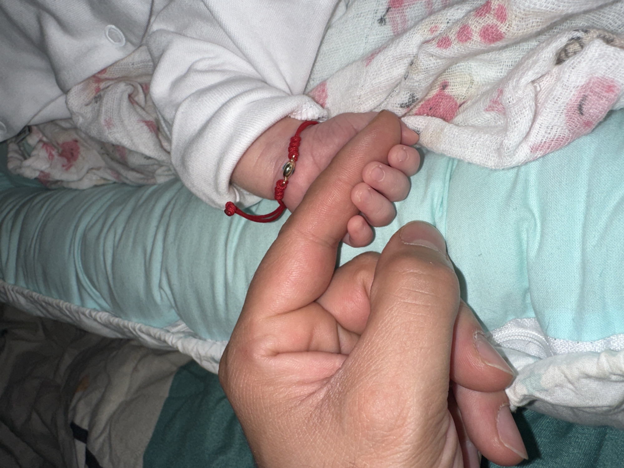 The image presents a close-up, intimate scene capturing the connection between a baby and an adult. The primary subjects are a baby's hand and an adult's hand. The baby's tiny right hand, adorned with a vibrant red string bracelet featuring a gold bead, is gently but firmly wrapped around the adult's index finger, which extends from the bottom right of the frame.
The baby is clad in a white long-sleeved garment, likely a onesie or top, and is surrounded by bedding. A soft, white fabric with a subtle pink pattern, possibly a blanket or swaddle, is visible around the baby's arm and hand. Below the baby, a light blue textured fabric, perhaps a fitted sheet or pillow, forms the background, while further down, a darker, patterned fabric suggests another layer of bedding.
The setting appears to be indoors, likely a bedroom or nursery, given the focus on hands and bedding. The activity depicted is a moment of connection, showing the baby's grasp on the adult's finger, which often symbolizes trust, comfort, and protection. The lighting is soft, creating a warm and peaceful atmosphere. There is no visible text in the image. The overall impression is one of tenderness and affection.