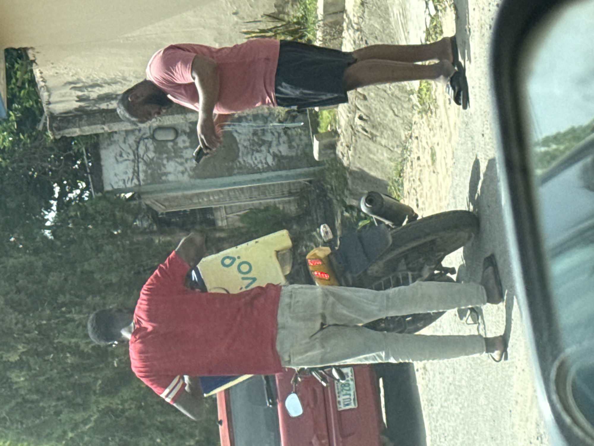The image depicts an outdoor street scene in what appears to be Abuja, Nigeria, oriented vertically. Two men are visible, standing near vehicles.
In the foreground, slightly to the left, a man wearing a red short-sleeved shirt and light-colored (possibly light grey or white) pants is standing with his back mostly to the viewer. He holds a rectangular yellow object, possibly a sign, in his left hand, on which the letters "V.O." are clearly visible. He is wearing dark sandals or flip-flops. To his right, a black motorcycle or moped is parked. Part of a red vehicle, possibly a car or truck, is visible below him, displaying a license plate with the registration "KUJ-28".
Further back and to the right, another man wears a pink short-sleeved shirt and dark shorts. He is looking down, seemingly at a dark object he holds in his hands, which appears to be a mobile phone. He is also wearing dark sandals or flip-flops.
The background shows sparse greenery, suggesting trees or bushes, and hints of light-colored buildings or walls. The scene is brightly lit, indicating daytime and sunny weather, with shadows cast on the ground. The men appear to be stationary, possibly waiting or engaged in a casual street-side activity, with the first man potentially advertising something with his sign. The image was captured from within another vehicle, as a side mirror is partially visible on the right edge.