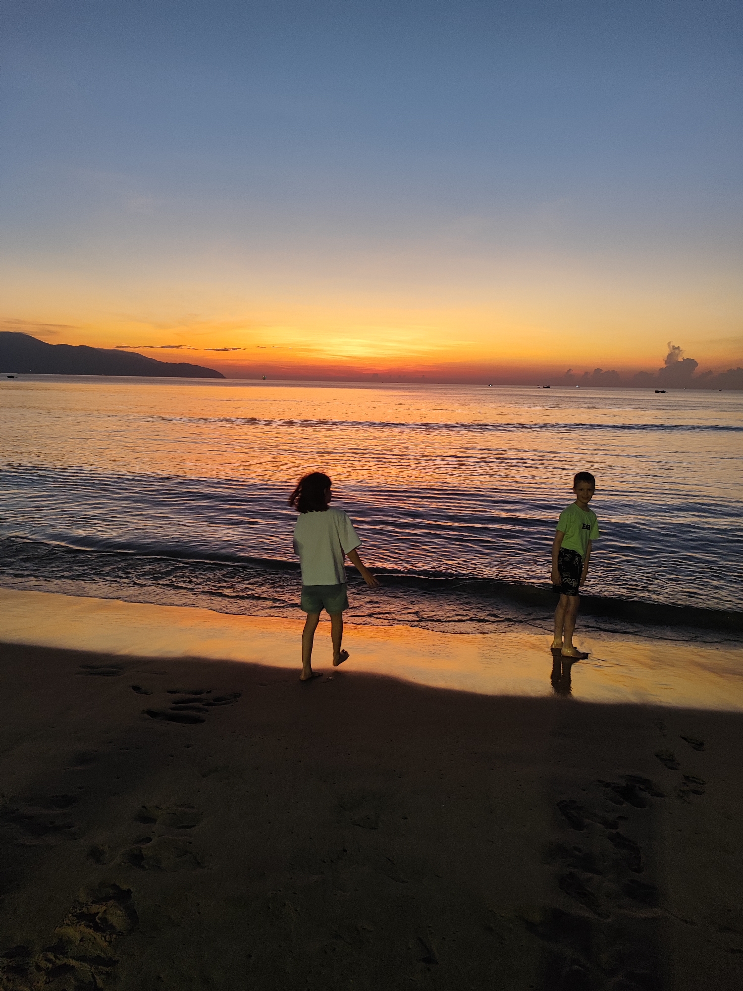 Two children are on a beach in Đà Nẵng, Vietnam at sunrise. One child with short dark hair is walking away from the viewer, wearing a white shirt and short dark shorts. The other child, a boy, stands to the right, facing the viewer. He has short brown hair and wears a lime green t-shirt and black shorts. The sun has just risen over the horizon, coloring the sky and water orange and yellow. The beach sand is dark. There are mountains in the distance. The water is calm. The children are partially silhouetted due to the bright light behind them. Footprints are visible in the sand.