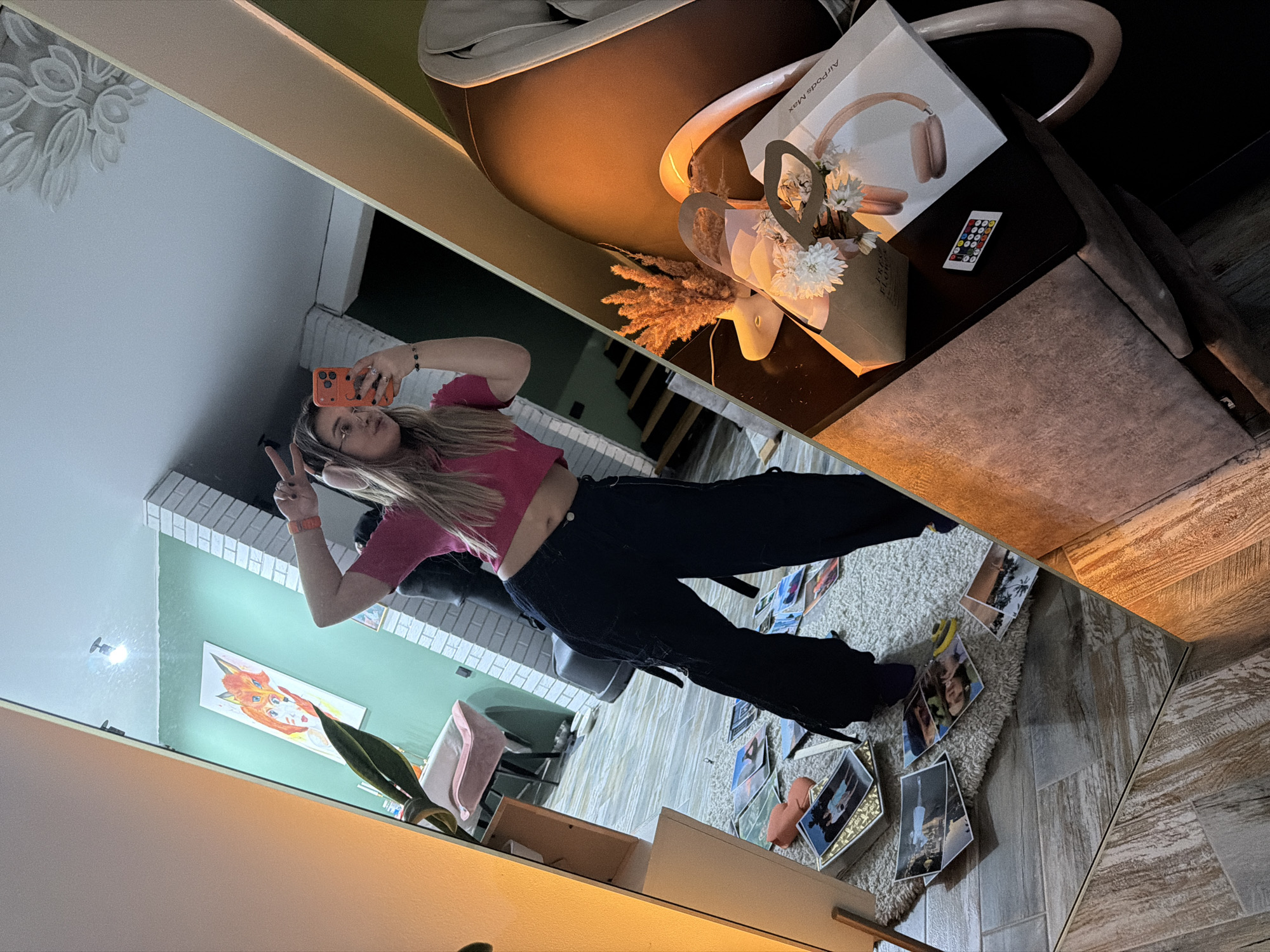 A mirror selfie shows a woman with long blonde hair, wearing a pink crop top and black pants, holding an orange phone and making a peace sign. The background includes a room with light green walls, a white brick wall, a fox painting, and a plant. Reflected on a table are a box labeled "AirPods Max," a bouquet of flowers, and a TV remote. A rug on the floor is covered in scattered photographs. The setting appears to be indoors, potentially in Большая Шильна, Russia, given the location provided.
