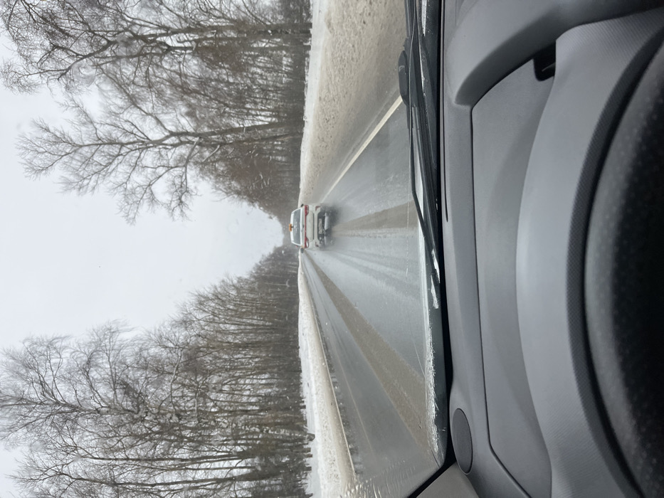 The image depicts a winter road scene in Podmosh'ye, Russia, viewed from the interior of a moving vehicle. The foreground shows the top of a dark gray dashboard and a portion of a steering wheel. Through the windshield, a two-lane asphalt road stretches into the distance, appearing wet with visible streaks of melting snow or slush. Snow-covered embankments and bare deciduous trees line both sides of the road. A white four-door sedan with an orange light mounted on its roof is visible approximately 20-30 meters ahead, driving in the same direction. Water spray is evident behind the sedan's rear tires. The sky is overcast. No people are explicitly visible outside the vehicles.