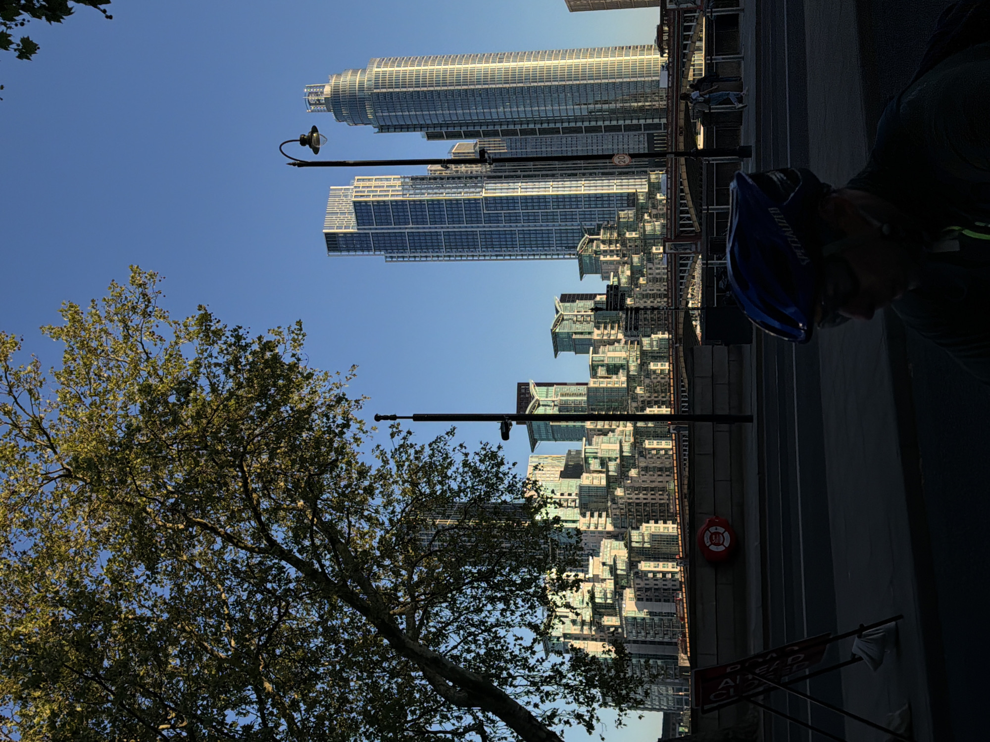 The image captures an urban landscape on a clear, sunny day, likely during late afternoon or early morning given the strong light and shadows. The scene is dominated by a clear blue sky, with a large, leafy tree prominent in the lower-left foreground, its branches extending upwards and across the frame.
In the background, a cluster of modern skyscrapers with reflective glass facades rises vertically. One notable building on the left features a distinctive cylindrical, ribbed design, while another next to it is a tall, rectangular structure. Beyond these, a dense array of smaller, multi-story buildings stretches into the distance, reflecting the sunlight.
On the right side of the image, a wide, dark paved area, possibly a street or promenade, recedes into the background. A street lamp extends from the right, reaching towards the center. A person, partially obscured and in shadow, is visible in the lower-right foreground, wearing a blue cycling helmet with the brand name "SPECIALIZED" clearly visible. Further back along the paved area, a red lifebuoy is attached to a structure, and a circular speed limit sign displaying "20" is mounted on a pole. Near the bottom right, a red sign with partially visible text reads "AHEAD CLOSED". The overall impression is of a vibrant city environment, consistent with London, United Kingdom.