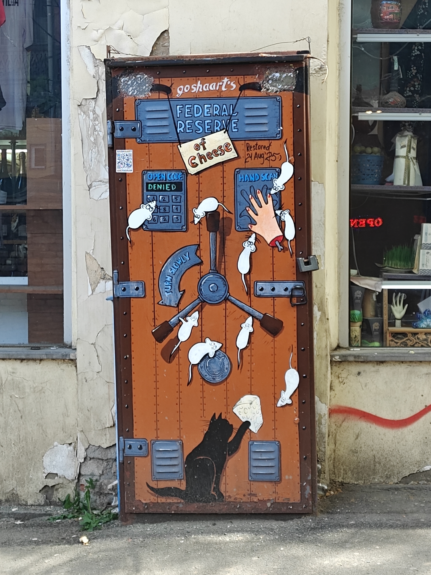 This image captures a vibrant and humorous piece of street art, or mural, painted on a metal door in an urban setting, likely Tbilisi, Georgia. The door itself is designed to resemble an old, rusted vault or safe, with rivets and a secure appearance.
The central subject of the mural is a satirical "goshaart's FEDERAL RESERVE of Cheese." Numerous white cartoon mice are depicted attempting to access this "reserve." One mouse interacts with a keypad displaying "OPEN CODE DENIED," while another is near a "HAND SCAN" panel, which features a cartoon severed hand. A large, mechanical vault handle is labeled "TURN SLOWLY." At the bottom of the door, a black cartoon cat holds a block of cheese, suggesting it has successfully pilfered from the "reserve."
Notable details include the artist's signature "goshaart's" at the top and a sign stating "Restored 24 Aug '25," playfully hinting at a future restoration. A QR code for "GOSHAART" is visible on the left side of the door. The surrounding environment features weathered walls with peeling paint and windows displaying various items, including partial text "TILIA" on the left and inverted "ИНЧО" (likely "OPEN" backward) on the right. A red spray-painted line curves across the wall to the right. The scene is brightly lit by daylight, suggesting a clear day. The entire artwork offers a whimsical and critical commentary on resources and access.