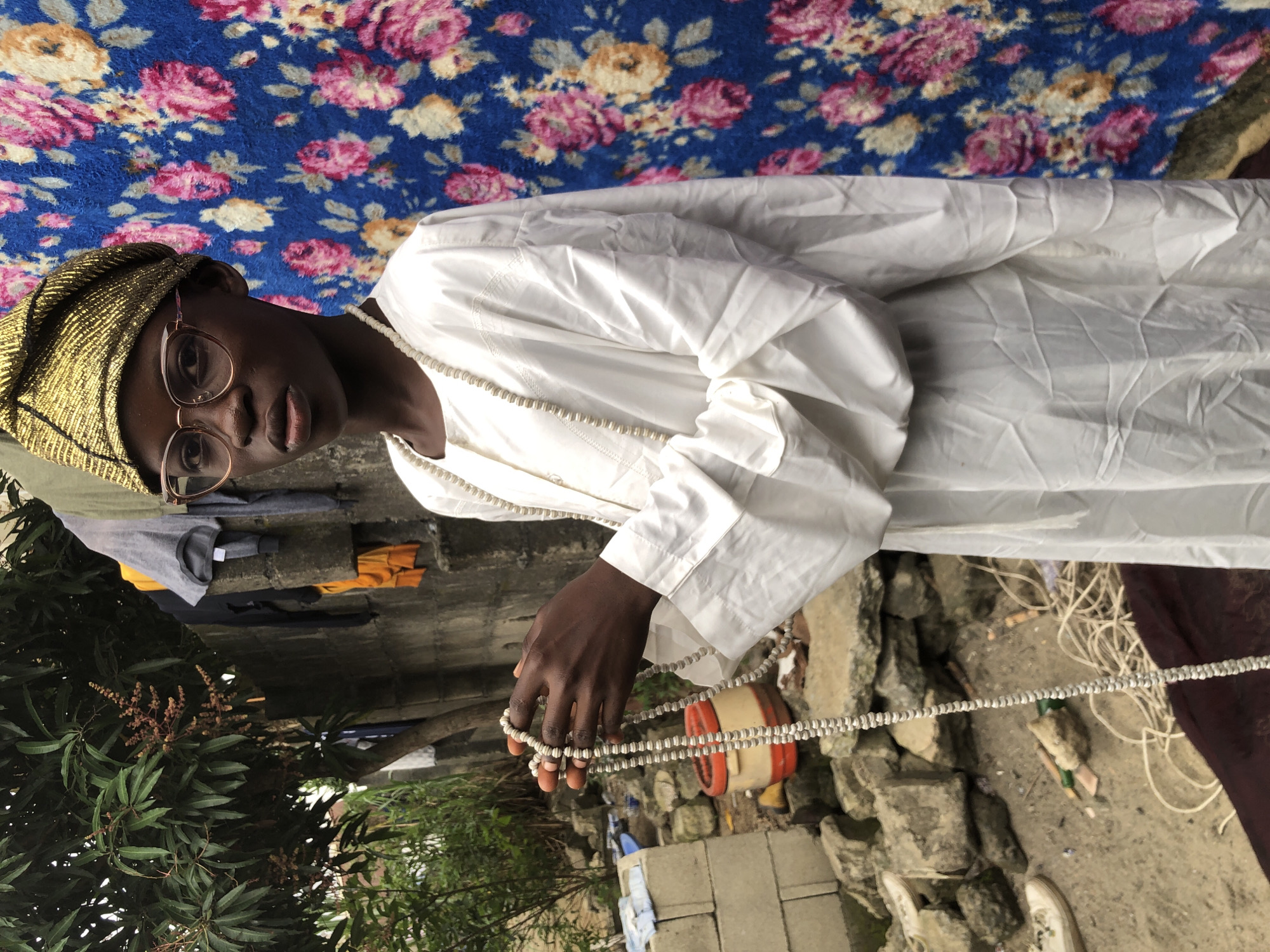 A young person wearing a white traditional robe, a gold headwrap, and glasses is standing outside. They are holding a long string of beads in their hands. The background features a concrete wall with clothes hanging on it, some greenery, rocks, and a blue cloth with a floral pattern. This scene is located in the city of Ado, Nigeria.