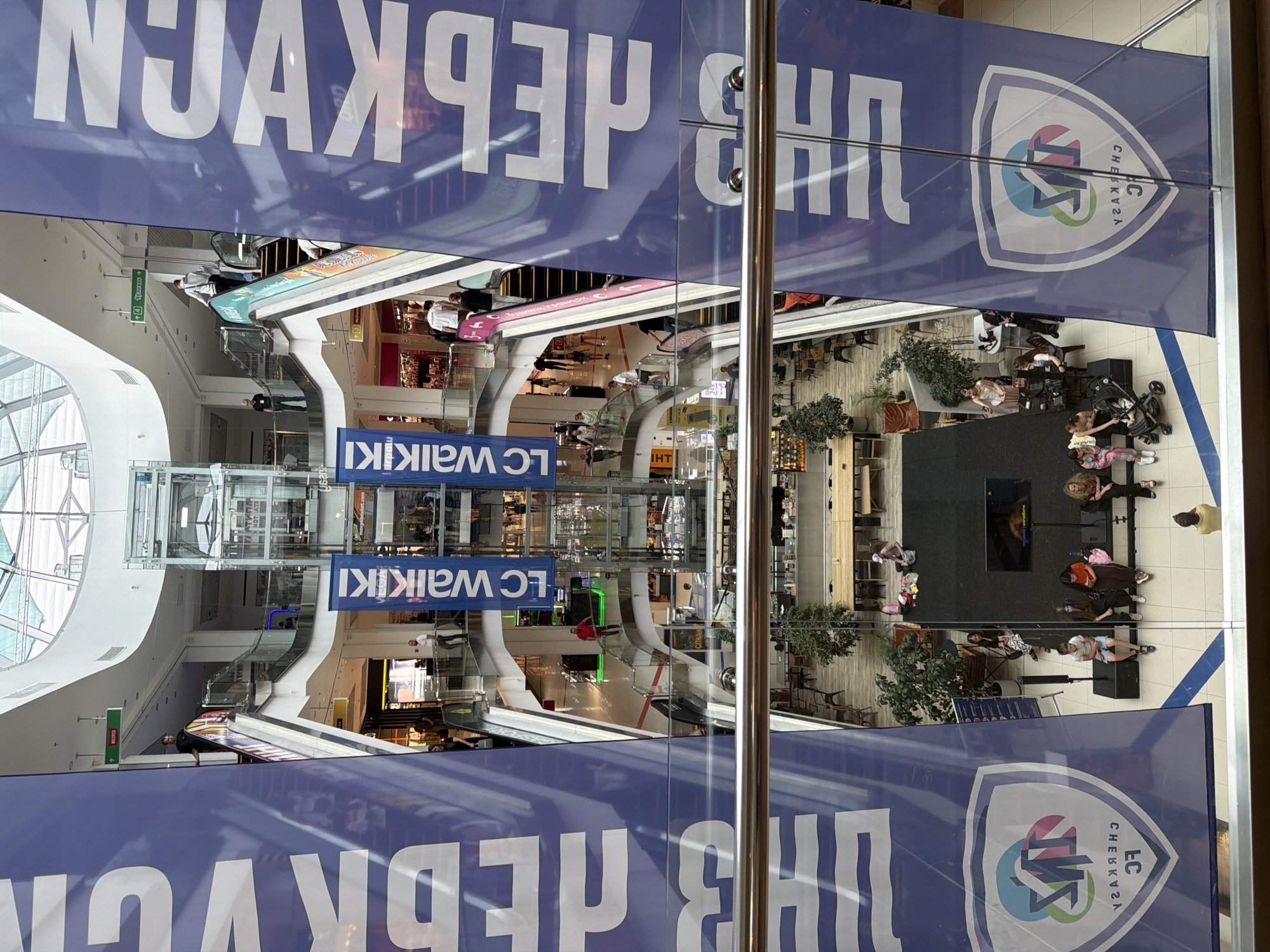 The image captures a multi-level interior of a shopping mall in Cherkasy, Ukraine. Dominating the foreground are two large blue banners with white lettering and a shield-shaped logo containing abstract shapes and text. The mall features multiple floors connected by escalators and an elevator shaft. People are visible on various levels, some walking, some standing, and some seated. On the lower level, there's a stage area with a screen and seating. The environment appears modern, with polished floors, glass railings, and bright lighting from the ceiling. Several shops and escalators are discernible on different floors. The overall scene depicts a busy public indoor space.