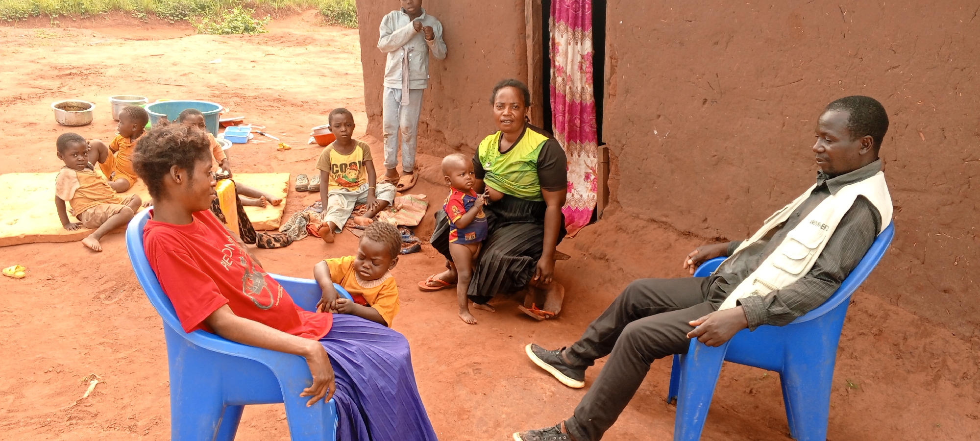 The image depicts a gathering of people, likely a family, outdoors in Makungu, Democratic Republic of the Congo. The setting is characterized by reddish-brown dirt ground and a simple building with unpainted, earthy-toned walls. A doorway, adorned with a pink and white floral curtain, is visible on the right side of the building.
In the foreground, a woman wearing a red t-shirt with "REDMON" partially visible text and a purple skirt sits in a blue plastic chair on the left. Beside her, a young child in an orange top is seated on the ground, looking towards the center. Further left, two other young children in orange tops are resting on a yellow mat, surrounded by metal pots and a blue plastic basin. A boy wearing a yellow t-shirt with "COOL LOVE" printed on it sits on the ground near the center.
On the right, a man in a black striped shirt and a light-colored vest, with "ANOTHER" visible on it, sits in another blue plastic chair, looking to his left. Between the man and the woman in red, another woman in a black and green top and a black pleated skirt is seated near the doorway. She is holding and breastfeeding a baby. A young boy in a light grey tracksuit stands against the building wall behind her.
The scene takes place during the day, under natural, soft lighting, suggesting either an overcast sky or indirect sunlight. The adults exhibit neutral to contemplative expressions, while the children are engaged in various activities or observing their surroundings. The image provides a glimpse into the daily life of a rural community.