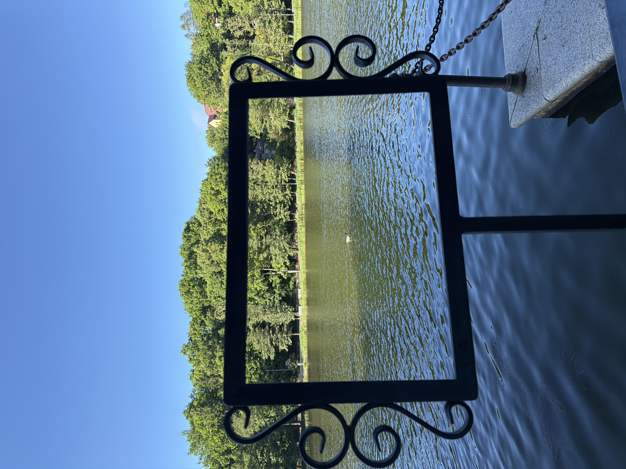 The image captures a serene lake scene in Svetlogorsk, Russia, bathed in the bright light of a clear day. The foreground is dominated by a decorative, black wrought-iron frame, possibly part of a signpost or a decorative element. This frame encloses a view of the lake, with its surface rippling under the sun.
Beyond the frame, the lake stretches out, its water reflecting the lush green of the surrounding trees and the bright blue of the sky. A solitary bird, possibly a duck or a goose, is visible in the distance on the water, adding a touch of life to the tranquil setting. The far bank of the lake is lined with a dense band of mature trees, their foliage a vibrant green. In the background, nestled amongst the trees, are glimpses of residential buildings with reddish-brown roofs, suggesting a settled area near the water.
To the right of the frame, a stone structure, likely a dock or a part of the lakeside infrastructure, is visible. A thick metal chain is attached to this structure, disappearing into the water, hinting at a mooring or a barrier. The overall atmosphere is peaceful and idyllic, evoking a sense of calm summer weather. The clear blue sky indicates daytime, likely mid-morning or afternoon, with no clouds present. There is no visible text in the image.