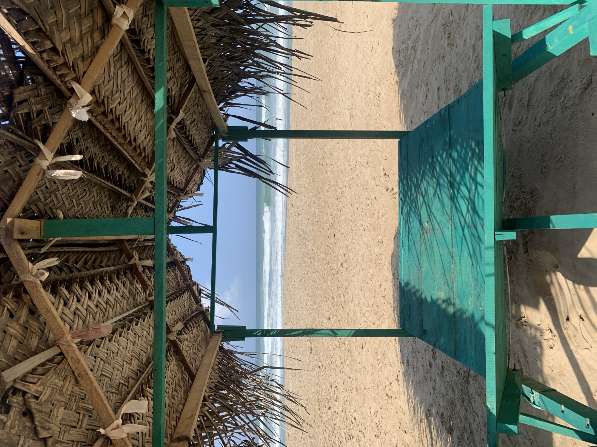 This image depicts a beach scene from Ahangama, Sri Lanka, as viewed through a green, wood-framed structure. The structure appears to be a platform or base with a thatched roof extending from the left. The roof is constructed of woven palm leaves and held together with wooden supports and white bindings. Beyond the structure, a sandy beach stretches out to the ocean, where gentle waves roll onto the shore under a clear sky. The lighting is bright, casting shadows from the structure onto the sand. There are no people visible in the shot.