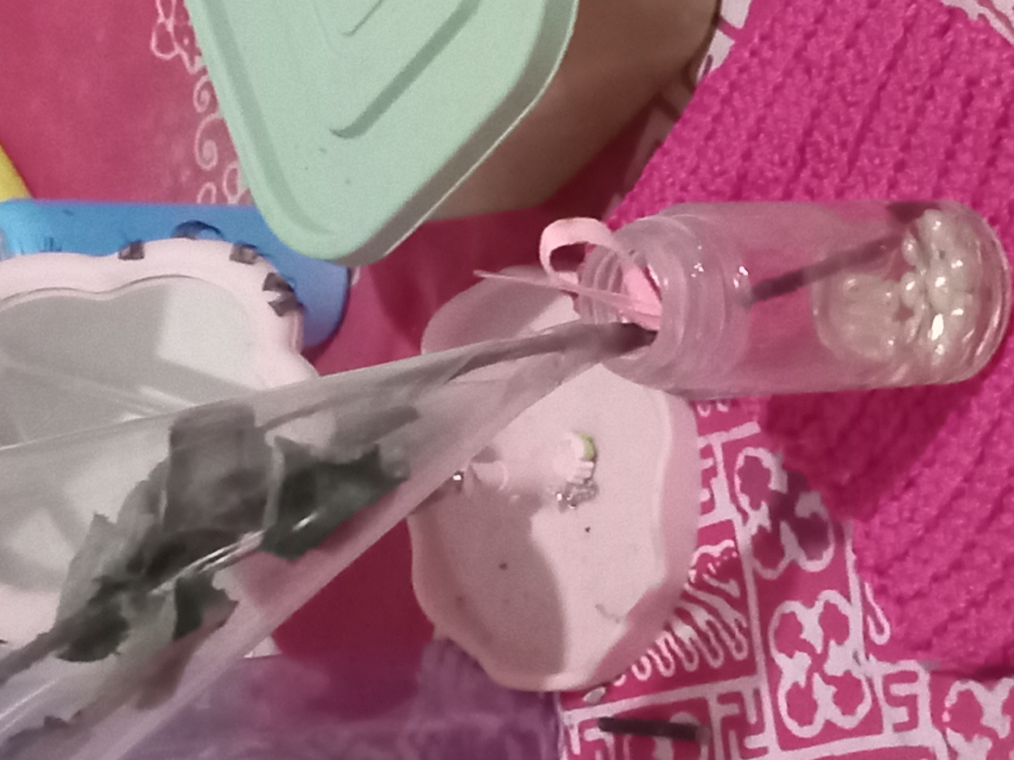 In Claket, Indonesia, an artificial green rose, encased in clear plastic, is being inserted into a small glass bottle containing white beads. The bottle has a pink ribbon tied around its neck and is placed on a pink knitted surface. To the left of the bottle, a pink heart-shaped dish with what appears to be a small silver charm is visible, situated on a patterned pink cloth. In the background, a blue cylindrical object and a light green lidded container are present, all on a pink surface. The lighting suggests an indoor setting.