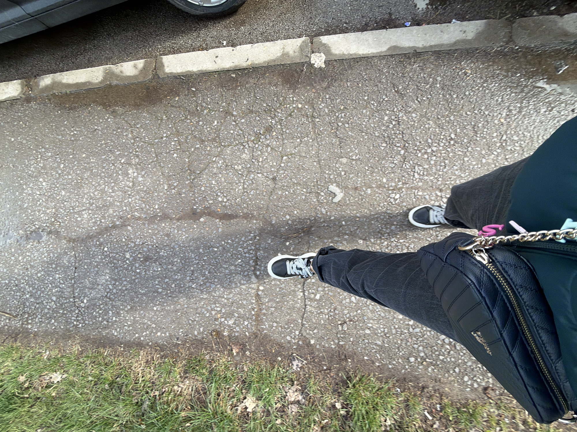 A first-person, overhead perspective image depicts a person standing on a cracked concrete sidewalk adjacent to a patch of grass and a curb. The person is wearing dark jeans and black sneakers with white laces. A black shoulder bag is visible, slung across their body. The sidewalk is gray and shows signs of wear and cracking. A curb runs along one edge of the sidewalk, and beyond that is a dark, indistinct area suggesting a road or parking area with part of a dark car visible. On the opposite side of the sidewalk, there is a strip of green grass. The location cannot be determined without additional information, but it is in an urban or suburban setting, possibly in Sofia, Bulgaria.
