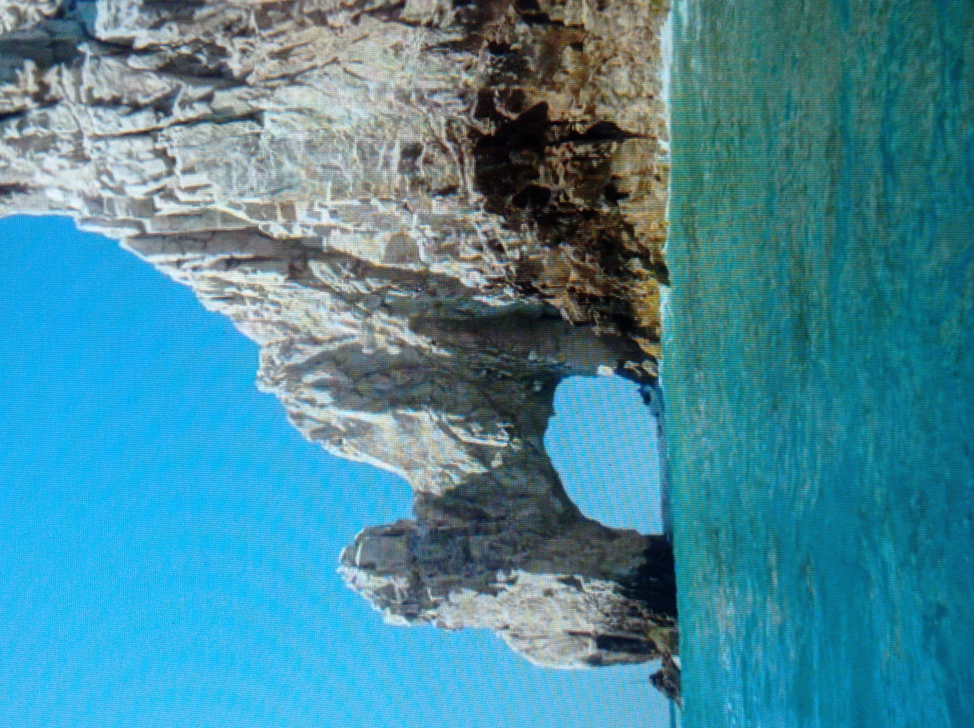 The image depicts a natural rock arch formation extending over the turquoise ocean under a clear blue sky. The rock is rugged and appears weathered, with dark shadows in some crevices. The water is calm, with some gentle ripples visible on the surface. The perspective is from the water, looking towards the arch. The location is not in Mukachevo, Ukraine.