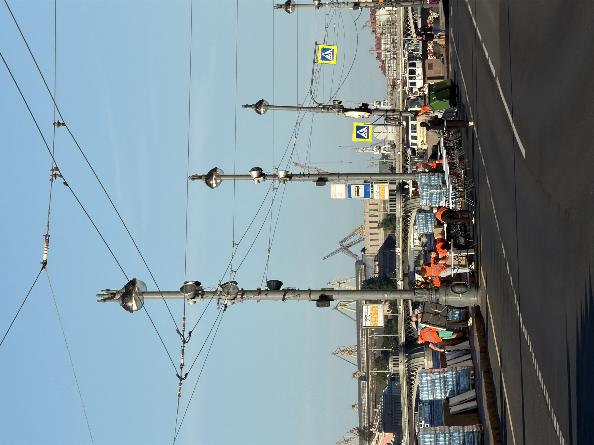 The image depicts a street scene in Saint Petersburg, Russia, likely on a bridge over the Neva River given the presence of a large body of water and the architectural style of the distant buildings and bridges. The foreground shows a road with white dashed lines. To the left, stacked pallets of bottled water are arranged alongside white boards. Several people, some wearing bright orange vests, are gathered around tables, appearing to be setting up or distributing supplies. They are engaged in various activities such as handing out items and organizing. In the background, a row of streetlights with ornate tops line the street. Further beyond, a cityscape unfolds with industrial cranes, buildings with red roofs, and a bridge crossing the water. Several boats are visible on the river. The sky is clear and blue, indicating a bright day. The overall atmosphere suggests an outdoor event or distribution point.