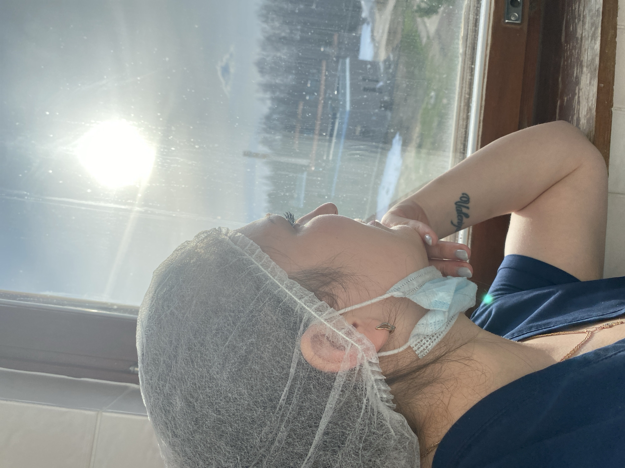 A woman wearing a light blue medical mask and hair net is reclining in front of a window. Her right arm is bent behind her head, revealing a tattoo on her inner forearm. She is wearing a dark blue garment and a gold chain necklace. The window shows a blurred, sunlit view of trees and buildings. The location is Марковский, Russia.