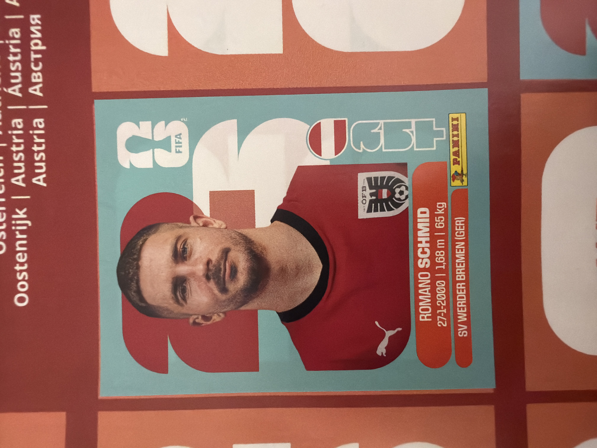 The image is a close-up of a sports trading card featuring a soccer player. The player, Romano Schmid, is shown from the shoulders up, wearing a red jersey with a black collar and the Puma logo. He has short, dark hair and a mustache. The card has an Austrian flag emblem on the right side of the player and a crest with the letters "ÖFB" and a soccer ball on the left. Text on the card includes "ROMANO SCHMID," his birth date "27-1-2000," height "1,68 m," weight "65 kg," and his club affiliation "SV WERDER BREMEN (GER)." A Panini logo is visible at the bottom right of the card. The background of the card features large, stylized numbers "23" in light blue and white. The overall image is part of a larger, likely printed material, with a red border surrounding the card and sections of the larger print visible. The location of Zlatitrap, Bulgaria is not depicted in this image.