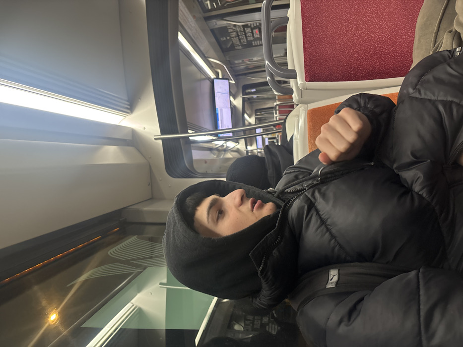 A person in a black puffy jacket and hood sits in a train car. The person's face is visible in profile, and their fist is clenched. Behind them, train seats are upholstered in red and orange. Above the seats, handrails are visible. The train car interior features a white wall with a window showing blurred scenery. Overhead lighting is present. Another passenger is sitting behind the main subject.