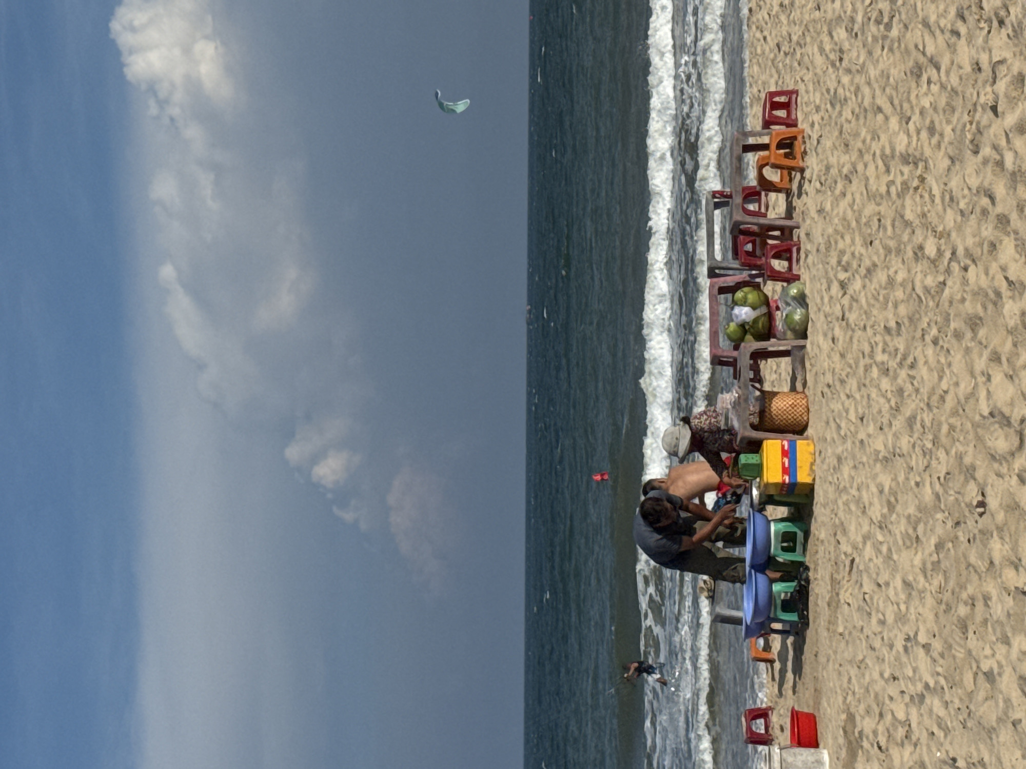 This image depicts a beach scene in Mũi Né, Vietnam. In the foreground, the sandy beach is occupied by a collection of low plastic tables and stools, suggesting a vendor stall. Several items are displayed on or around these tables, including what appear to be coconuts and some wrapped goods. Two people are actively engaged at the stall, one leaning over the items, and another seated. Further in the background, the ocean is visible with waves crashing onto the shore. A person is seen kiteboarding in the water to the left, and a kite is flying in the sky to the right. The sky is partly cloudy, with a mix of blue sky and white clouds. The overall atmosphere is that of a typical sunny day at a Vietnamese beach.