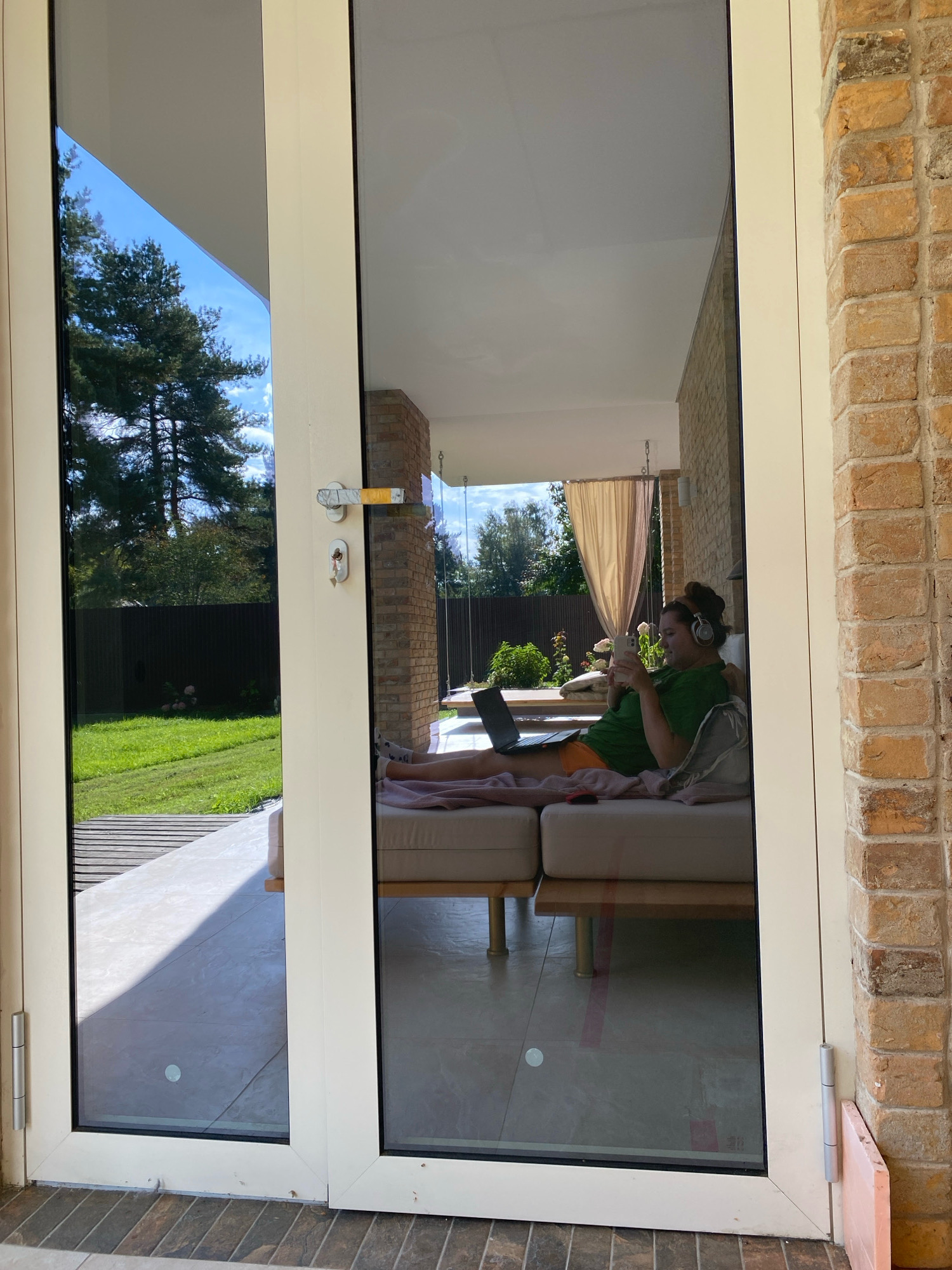 A woman is seen through a glass door, relaxing on a sofa with a laptop and a phone in front of her. She is wearing headphones and appears to be engaged with her devices. The reflection in the glass shows a backyard with trees, a fence, and a grassy area. The exterior of the building is made of brick. The scene is well-lit, suggesting it is daytime. There is no information in the image that would allow for the determination of the specific location within the city of Perm, Russia.