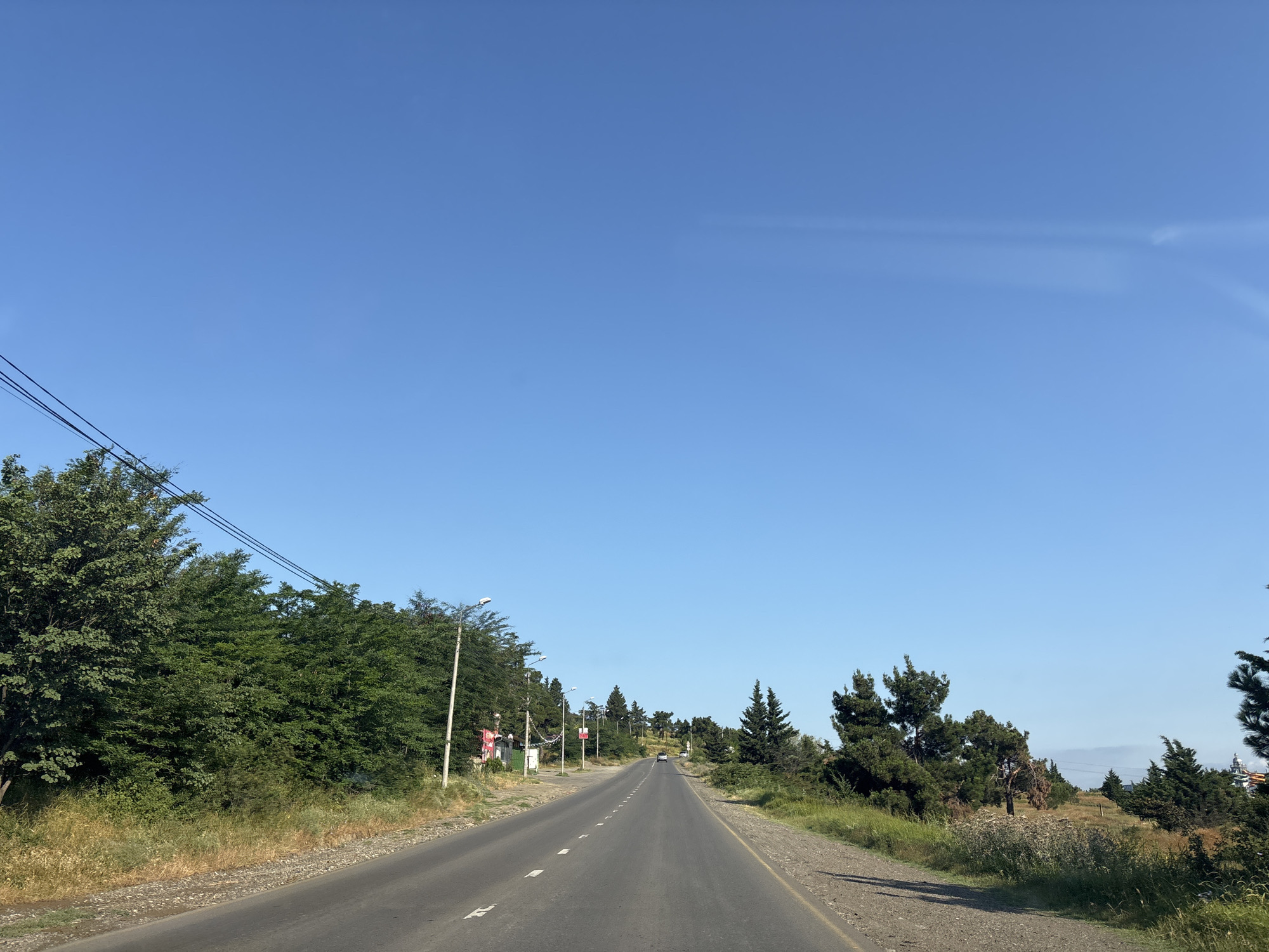 The image depicts a straight, paved road in a rural or suburban area of Tbilisi, Georgia, under a clear blue sky. The road has white dashed lines marking the lanes and a yellow line on the right edge. Dense green trees and vegetation line both sides of the road, with some scattered evergreen trees further into the landscape. There are utility poles and wires visible on the left side, along with some small structures or buildings. A few cars are seen in the distance on the road. The overall impression is of a sunny day in a somewhat undeveloped area outside the main city center.