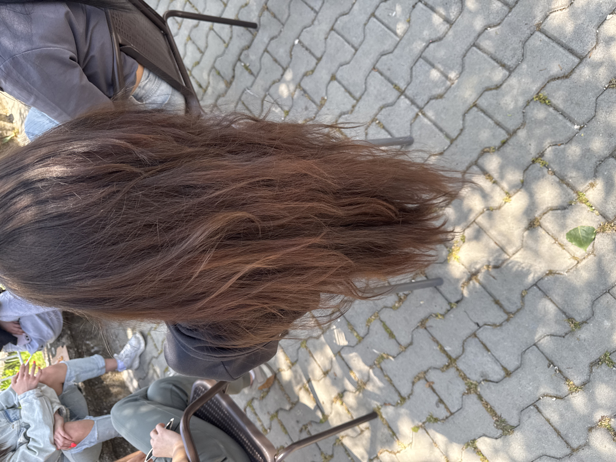 The image shows a close-up, top-down view of a person with long, light brown hair. The hair is spread out on a paved surface with interlocking brick patterns. The person is sitting in a metal chair. Portions of other people sitting nearby are visible in the lower left corner, wearing jeans and jackets. The location appears to be an outdoor area with stone pavement.