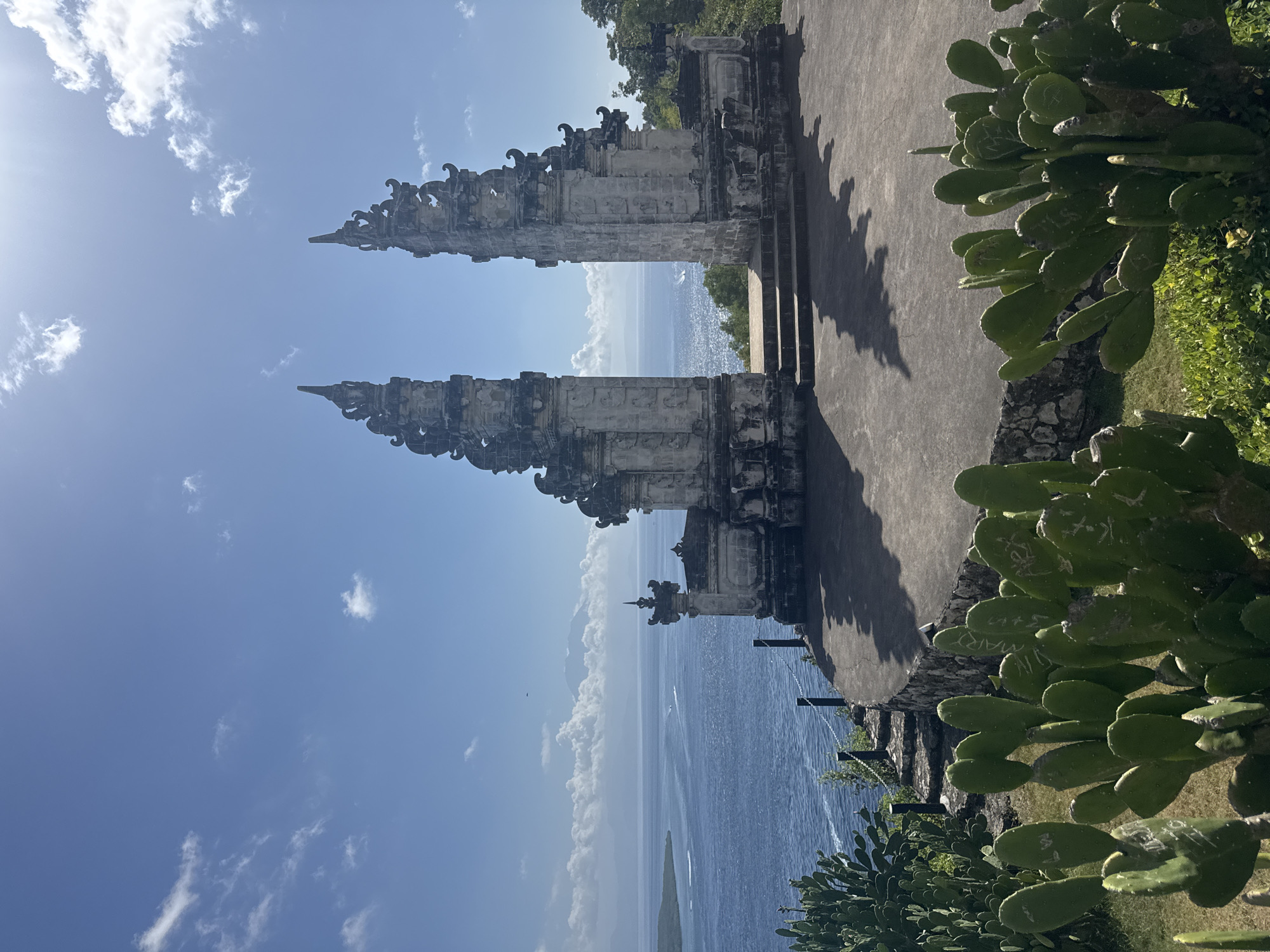 An outdoor, medium-shot, eye-level view captures the Pura Lempuyang temple gates on the island of Nusa Penida, Indonesia. The sky, a vibrant blue, is partially obscured by scattered white clouds and the bright glare of the sun. The temple gates, constructed of weathered stone with intricate carvings, stand prominently in the center of the frame. They are open, revealing a breathtaking vista of the ocean and distant mountains. The ocean, a deep blue, is dotted with small boats and reflects the sunlight. In the foreground, lush green cacti, some with carved inscriptions, thrive in the arid environment. To the left of the frame, stone steps lead down a hillside, bordered by a stone wall and a chain-link fence. The ground in front of the temple is a large, flat stone patio with dark shadows cast by the sun. The overall scene evokes a sense of awe and natural beauty.