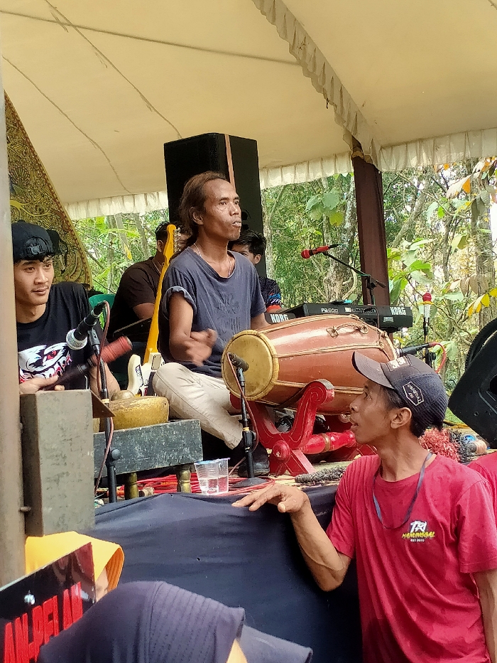 This image depicts an outdoor musical performance or rehearsal under a light-colored tent canopy in Tamansari, Indonesia.
In the center, a male musician, appearing middle-aged with long dark hair, sits cross-legged. He wears a dark blue t-shirt and light-colored trousers, and his hands are positioned over a large, reddish-brown kendang drum on a red wooden stand. A microphone is situated near the drum. Behind him to the right, a large black speaker cabinet is visible, and further back are two black electronic keyboards, one labeled "KORG" and another "Pa500," with an additional microphone on a stand.
To the left of the central drummer, a younger male musician, wearing a black t-shirt with a white graphic and a backward black baseball cap, sits. He holds a mallet over a rectangular wooden frame containing a small metal percussion instrument, possibly part of a gamelan ensemble. A microphone is also placed near this instrument. A patterned brown and gold fabric panel hangs vertically from a tent support pole to his left.
In the foreground on the right, another male individual, wearing a red t-shirt with a "TA HOMERGAL EST 2020" logo and a black baseball cap worn forward, leans forward. His right hand rests on a black-draped surface, and he looks up and to the left, observing the scene.
The performance area is draped in black fabric, with cables and equipment visible on the surface. A clear plastic cup containing liquid sits near the kendang drum. In the background, beyond the tent structure, lush green foliage and tree branches are visible, indicating an outdoor setting. Several other blurred figures are present in the background, including one behind the central drummer with a yellow electric guitar visible.