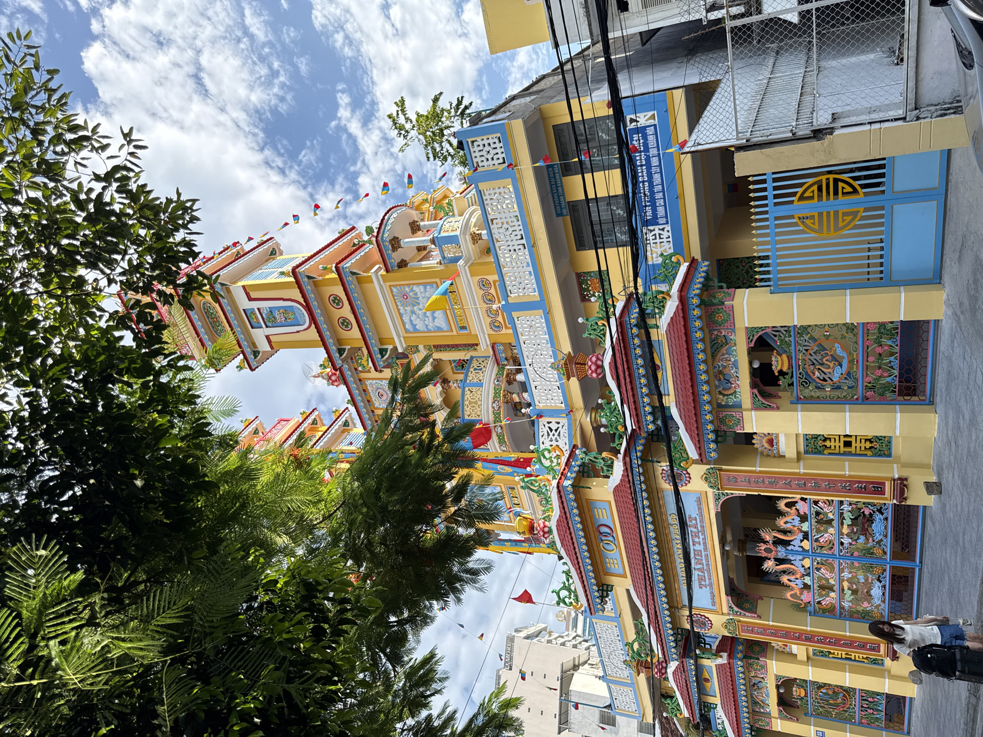 This is a full, eye-level, outdoor, daytime shot of the ornate, yellow and blue facade of a temple in Nha Trang, Vietnam. The temple has multiple levels, decorative roofs, and colorful banners and flags. The architecture is elaborate with intricate carvings and statues. In the foreground, a young woman wearing a white t-shirt and denim shorts stands with a black backpack near the entrance. To the right of the temple is a blue gate with a yellow symbol. Overhead, lush green foliage partially obscures the top of the temple and the sky, which is dotted with white clouds. The image captures a vibrant, culturally rich scene in the city.