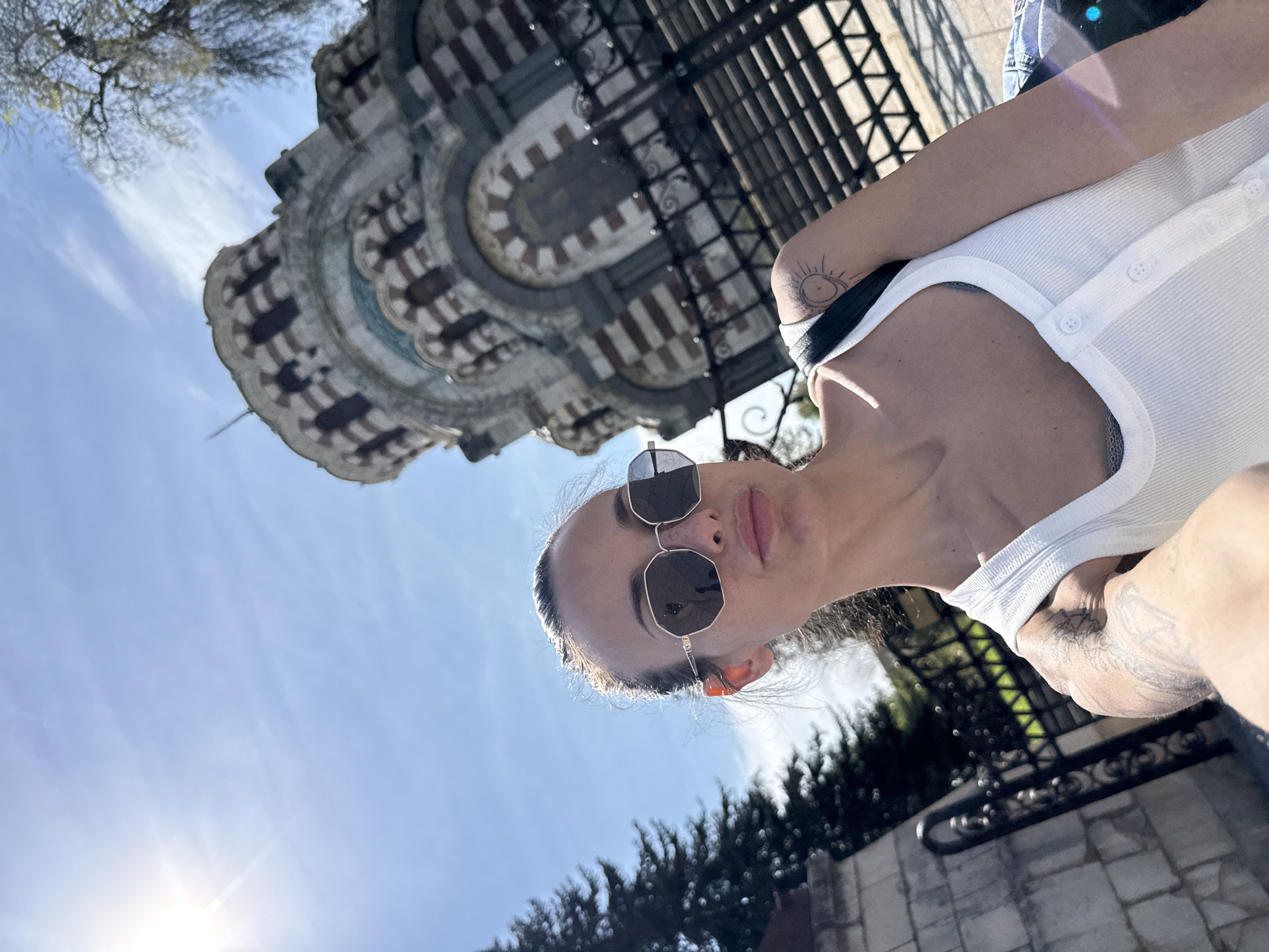 An upward-angled shot captures a woman in sunglasses against a historic building and a bright sky, likely taken in Pleven, Bulgaria.
The primary subject is a woman, positioned towards the right of the frame. She wears a white ribbed tank top and dark, geometric-framed sunglasses, which reflect some of the surrounding light. Her dark hair is pulled back, catching highlights from the sun positioned behind her. Visible details include a small sun-like tattoo on her right arm and a larger, intricate tattoo, possibly a wing or detailed design, on her left inner arm/shoulder area. Her expression is obscured by the sunglasses, but she appears relaxed, looking towards the camera.
In the background, dominating the left side of the frame, stands a large, ornate architectural structure. This building strongly resembles the Mausoleum Chapel "St. George the Conqueror" in Pleven, characterized by its distinctive horizontal bands of light and dark stone or brickwork, forming a striped or patterned facade with arched window or decorative elements.
The scene is set outdoors during daytime, indicated by the clear blue sky with some wispy clouds. A bright sunburst is visible towards the lower left of the frame (when oriented correctly), casting strong light. A black metal fence or gate is visible behind the woman and to the right, with glimpses of green foliage from trees in the distance. No visible text is present in the image.