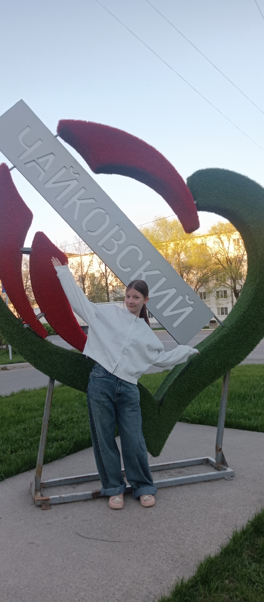 A girl stands in front of a large heart-shaped sign in the city of Unknown, Unknown. The sign is made of red and green material with a gray banner across it that reads "Чайковский" in Cyrillic script. The girl is wearing a light gray zip-up jacket, blue jeans, and pink shoes. She has her arms outstretched, and she's smiling at the camera. The background includes trees, buildings, grass, and a paved area. The sky is light blue, indicating it is either early morning or late afternoon.