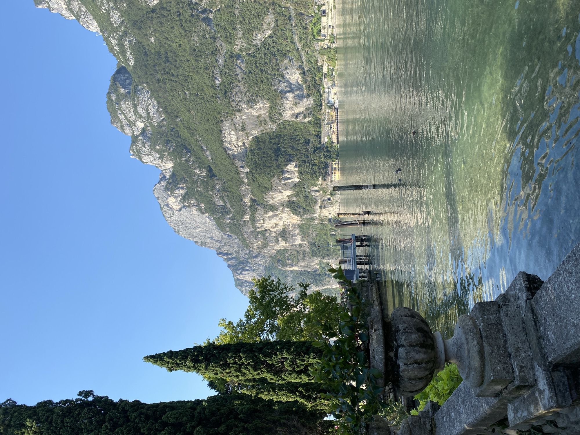 The image depicts a serene lakeside scene, likely in Riva del Garda, Italy, given the distinctive mountainous backdrop. In the foreground, a stone balustrade and a decorative urn, laden with greenery, frame the view. Beyond this, a tranquil body of water, the lake, reflects the clear blue sky and the imposing, tree-covered mountains that rise steeply in the background. The water is remarkably clear, revealing the lakebed in some areas. Several wooden posts rise from the water, suggesting a dock or mooring area. A modern pier extends into the lake, with its railing visible. In the distance, along the lakeshore, a cluster of buildings, possibly hotels or residences, are nestled at the base of the mountains. Two ducks are visible swimming on the surface of the lake, adding a touch of life to the picturesque setting. The overall impression is one of peaceful natural beauty, characteristic of the Italian Alps.