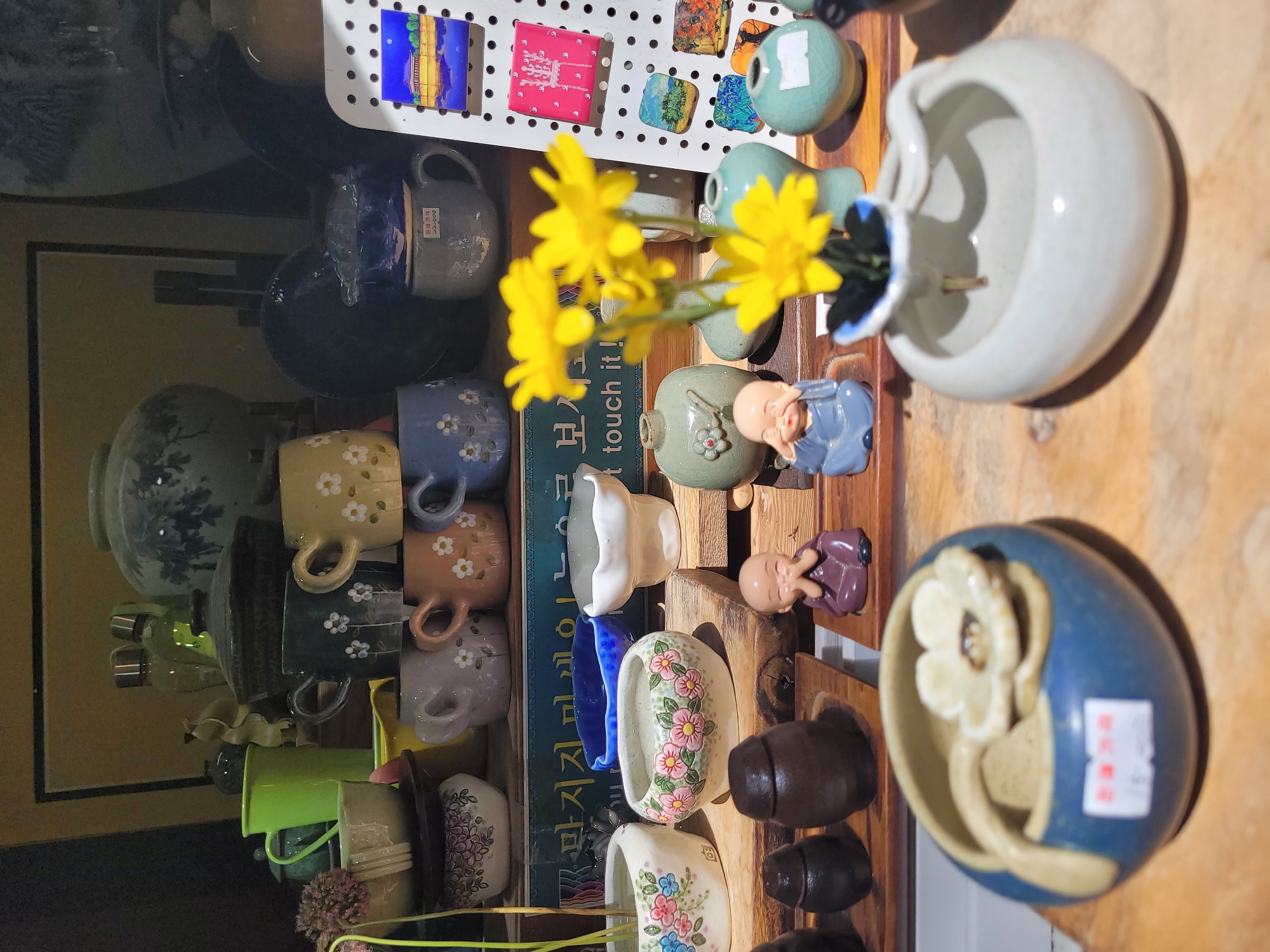 A shelf in a shop in Gyeongju-si, South Korea, displays a collection of ceramic items, primarily small pots and cups, alongside decorative figurines and a few yellow flowers in a vase. The collection is arranged on wooden shelves. In the foreground, a blue ceramic pot with a white flower ornament and a spoon sits on a wooden plank. To its right, a white ceramic vase holds a stem with a yellow flower. Behind these, a row of small, earth-toned jars and two monk figurines are visible. Further back, various ceramic cups with floral patterns in shades of green, blue, grey, and brown are stacked. There are also some larger ceramic vases and decorative plates on higher shelves. The overall impression is of a display of handcrafted ceramics, likely for sale.