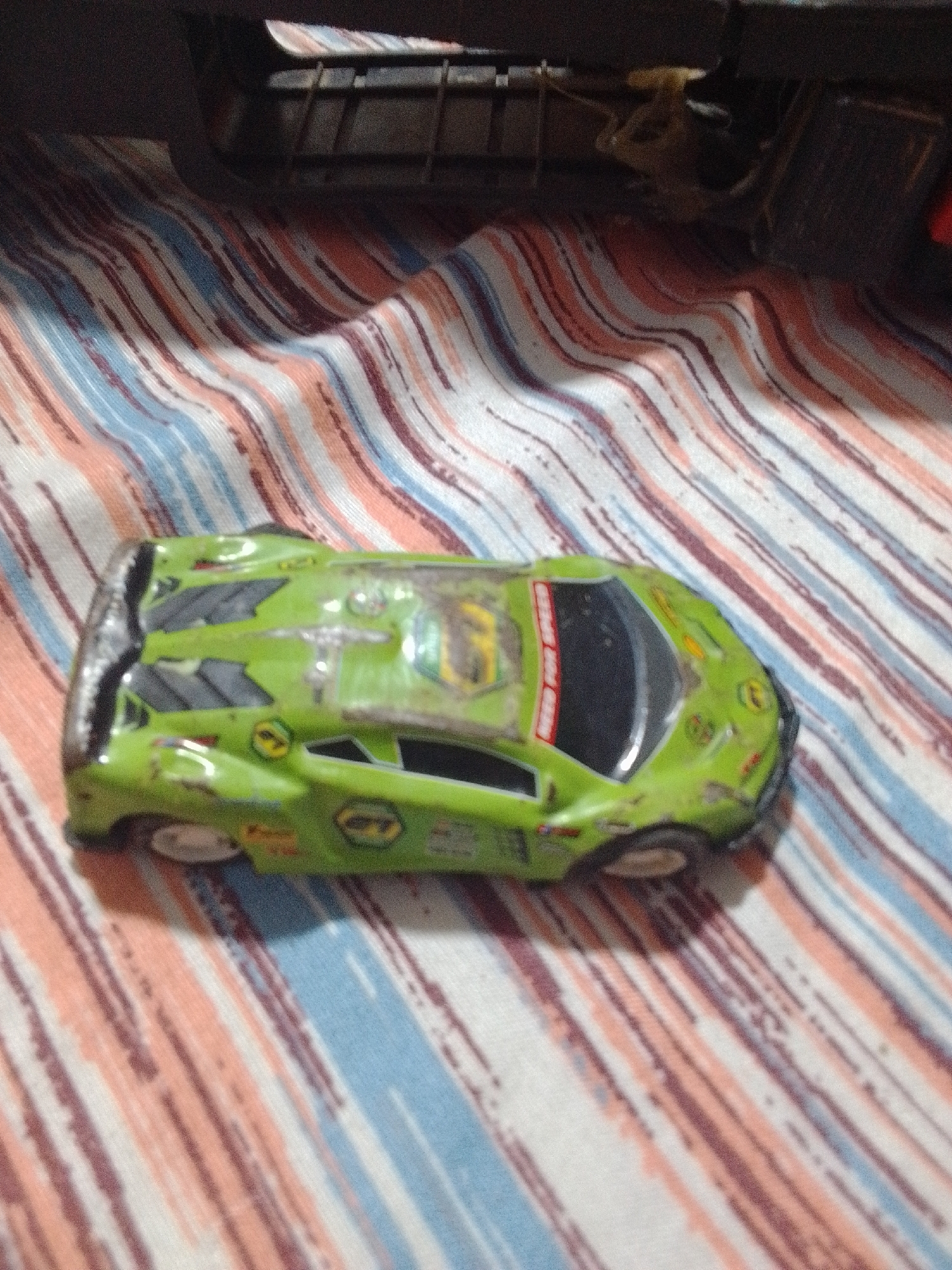 An overhead shot shows a green toy car with a clear top resting on a striped fabric surface. The car has black detailing on its sides and a red stripe on its roof. The fabric has diagonal stripes of white, light blue, and a reddish-brown color. The background is dark and out of focus, suggesting it is a table or shelf. There is no indication of the location being in La Tagua, Venezuela.