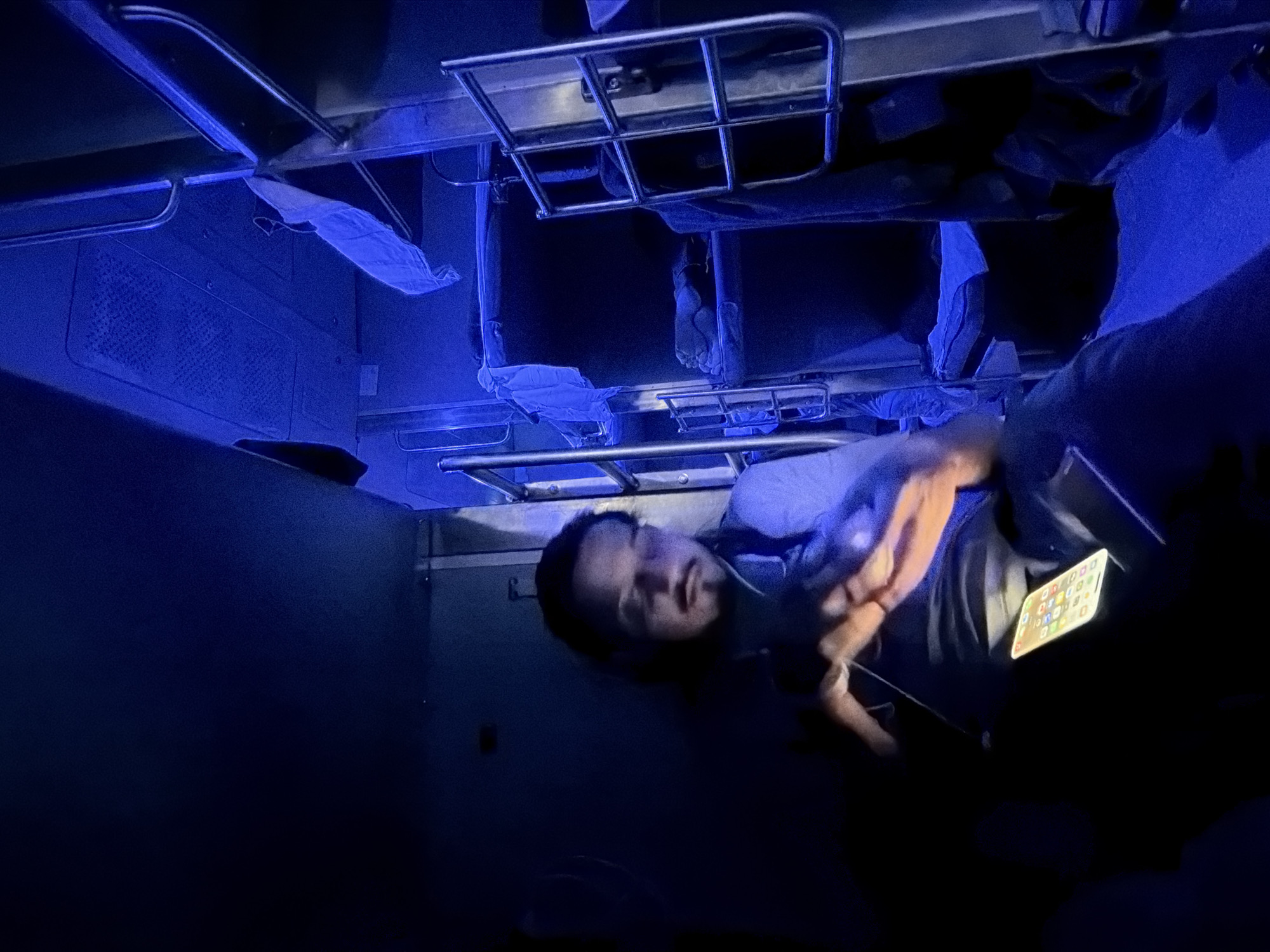 The image shows a man sitting in a dimly lit train compartment, illuminated by blue lights. He is wearing headphones and looking at his phone, which is resting on his lap. The compartment has bunk beds with blue blankets and white sheets. The man has a mustache and goatee and appears to be in his late twenties or early thirties. The scene suggests a journey on a train, possibly at night, given the darkness and the artificial lighting. The location is identified as Malya (Alot), India, a town in the Ratlam district of Madhya Pradesh, known for its railway station, which could explain the train compartment setting.