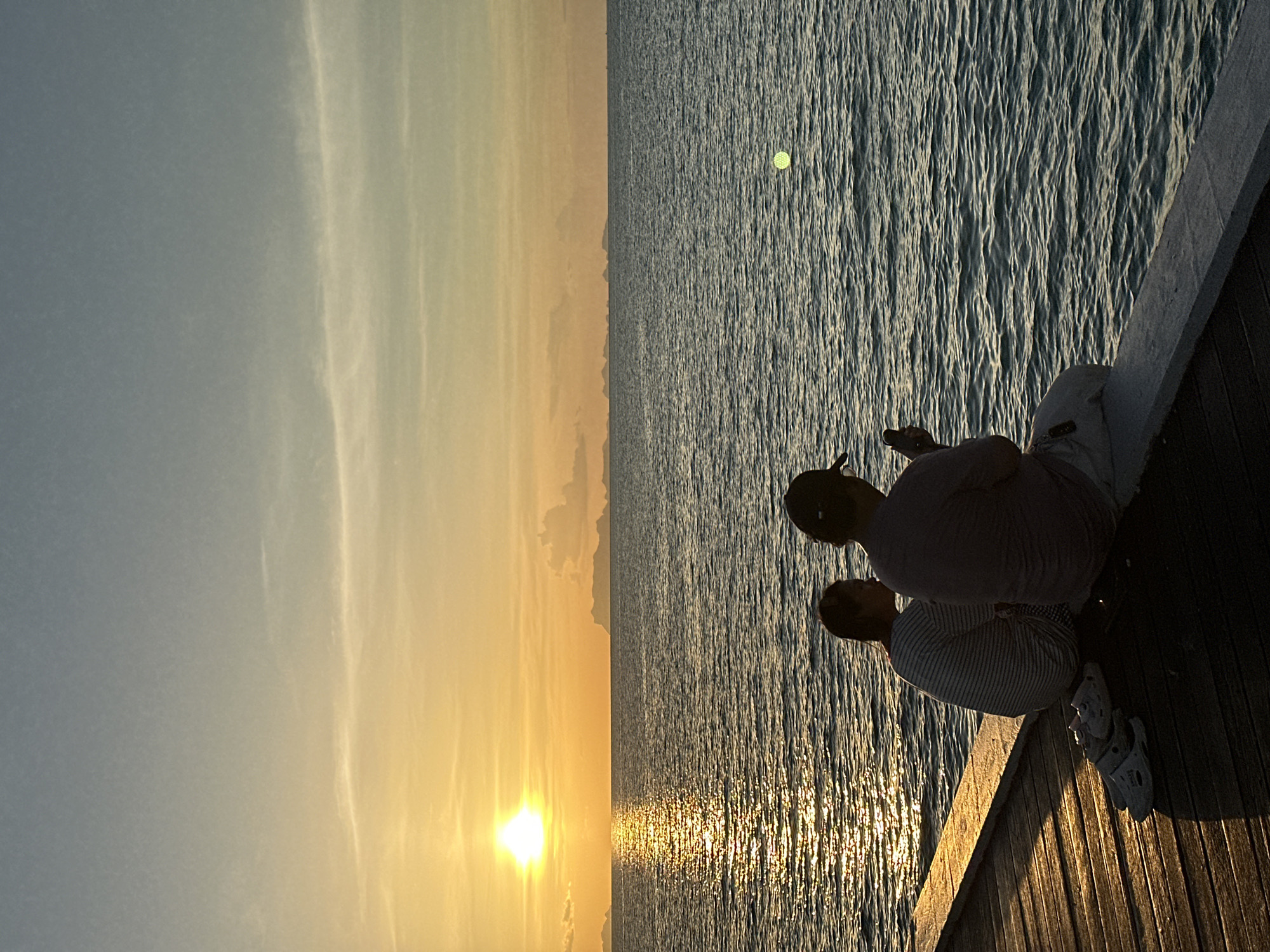 Two individuals, silhouetted against a vibrant sunset, sit on a wooden pier overlooking a tranquil ocean. The person on the left, a younger individual, is facing away from the camera, while the person on the right, an adult wearing a cap, is looking down at a mobile phone. Scattered on the wooden planks of the pier are a pair of white slip-on shoes. The setting sun casts a golden hue across the water, creating a shimmering reflection. In the distance, a faint outline of islands can be seen against the horizon. The sky transitions from a bright orange near the sun to a soft blue at the top. The overall scene suggests a peaceful evening at the beach in Baan Taling Ngam, Thailand.