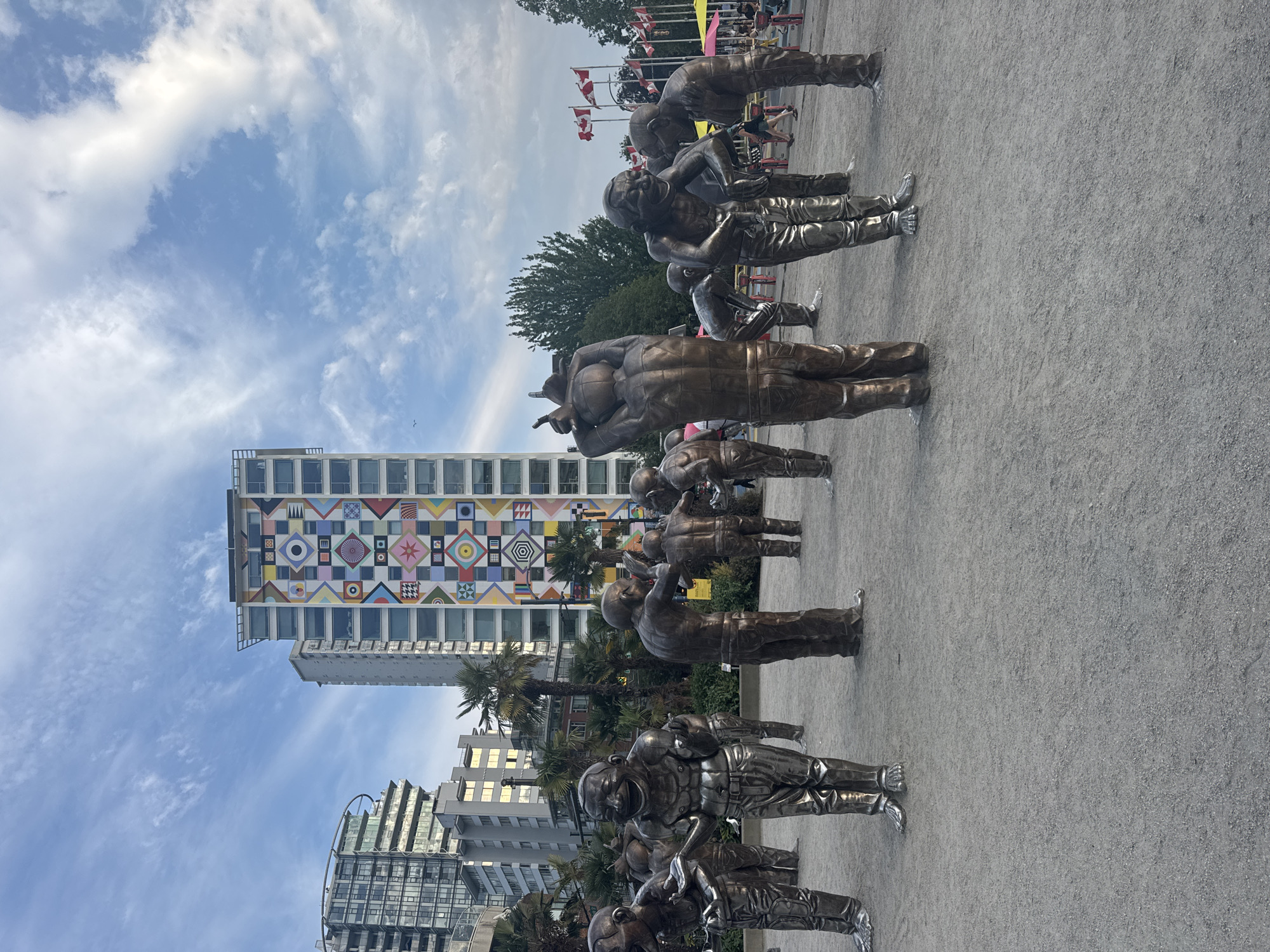 The image depicts a public art installation of bronze laughing figures in a plaza in Vancouver, Canada. The sculptures are arranged in a loose cluster, with some figures appearing to interact with each other through gestures and posture. The ground is paved with gravel, and palm trees are visible in the background, suggesting a temperate climate. Behind the sculptures, modern high-rise buildings dominate the skyline. One building in particular stands out with a vibrant, geometric mural on its facade. The sky is a mixture of blue with scattered white clouds. Several flags are also visible in the distance, along with some people walking in the background.