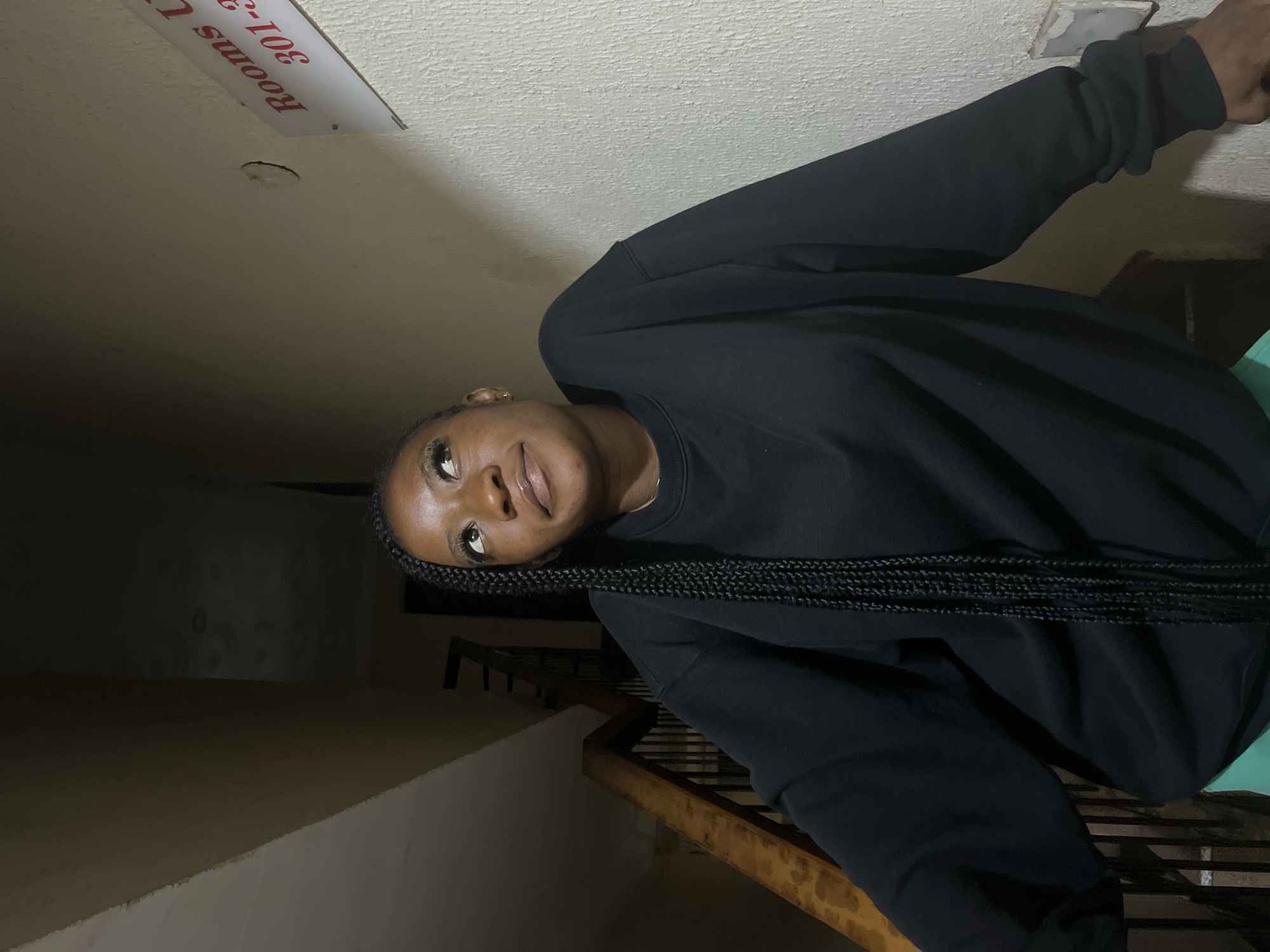 A medium shot captures a young Black woman with long braided hair, wearing a black sweatshirt and green shorts. She is leaning against a wooden handrail on a staircase. Her head is tilted upwards, and she gazes to her right with a slight smile. To her right, on a textured wall, a sign reads "Rooms U" with "301-3" visible below. The lighting is dim, with shadows indicating an indoor setting, likely a stairwell. The image is set in Jos, Nigeria, a city known for its elevated plateau and cooler climate, often featuring multi-story buildings with concrete and wooden staircases.