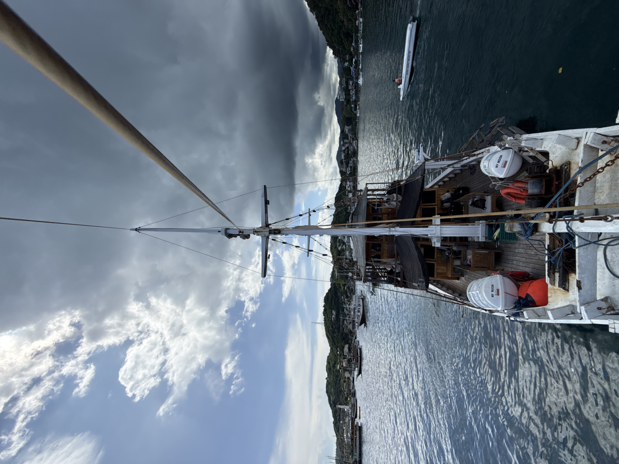 The image depicts a view from the deck of a boat docked in Labuan Bajo, Indonesia. A sailboat mast extends upwards towards the sky, which is divided between dark, ominous storm clouds and patches of blue sky with white clouds. The water is relatively calm with reflections of the sky and surrounding landscape. On the water, a white vessel with a person visible is seen. The boat has a weathered wooden deck and white painted elements. Life preservers and equipment are visible on deck. The shoreline shows green hills with buildings clustered along the coast. Other docked boats can be seen along the shoreline.