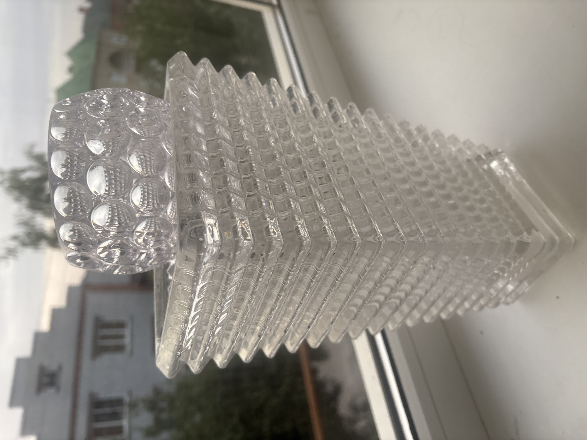 The image displays a clear glass vase with a stepped, square base and a textured cylindrical top. The vase is positioned on a white windowsill against a blurred background of buildings and trees, likely in Georgiyevsk, Russia. The architecture visible in the background includes a building with a stepped roofline and another structure with a green roof, consistent with urban or suburban settings. The lighting suggests it is daytime, with overcast skies. No people or actions are depicted.