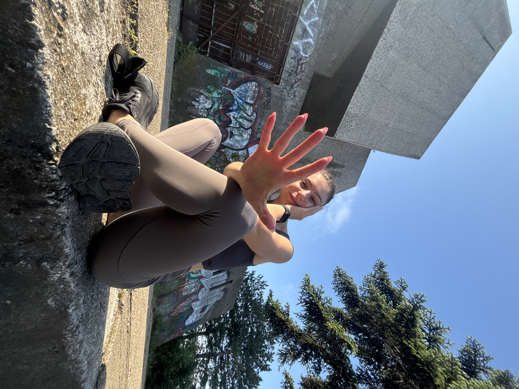 This media file presents a dynamically angled, low-perspective portrait of a young woman resting outdoors in Varna, Bulgaria, on a clear, sunny day. The subject, a woman with dark hair pulled back, is wearing dark grey/brown athletic leggings and a matching sports bra or top, along with black athletic shoes. She has bright pink nail polish and a black smartwatch on her left wrist.
She is depicted lying or leaning on a rough concrete surface, with her legs bent and one foot's sole facing the camera. Her right hand is raised, palm open, playfully covering part of her smiling face, while her left hand gently rests against her jaw. Her expression is cheerful and relaxed.
The setting combines urban elements with natural scenery. Behind the woman, a concrete wall is heavily covered in colorful graffiti, featuring various abstract shapes and some distinct Cyrillic-style lettering, including text that appears to be "PABXA" and "ODOL". Above this wall, a large, angular concrete structure, possibly a Brutalist building or monument, is seen from below, presenting a textured, weathered surface against the expansive blue sky. To the lower right, verdant coniferous trees rise into the frame, adding a touch of nature. The bright sunshine indicates it is daytime. The unique angle gives the scene a dynamic and somewhat artistic feel.